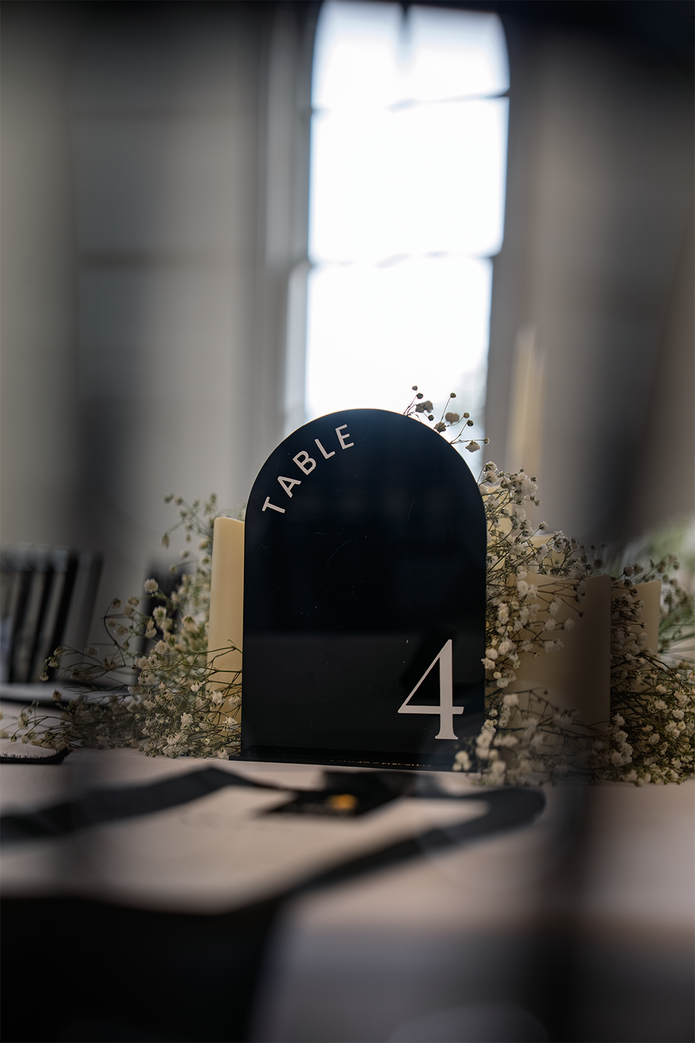 Table number 4 at a formal event, with a black table number sign surrounded by white flowers, in front of a window with natural light.