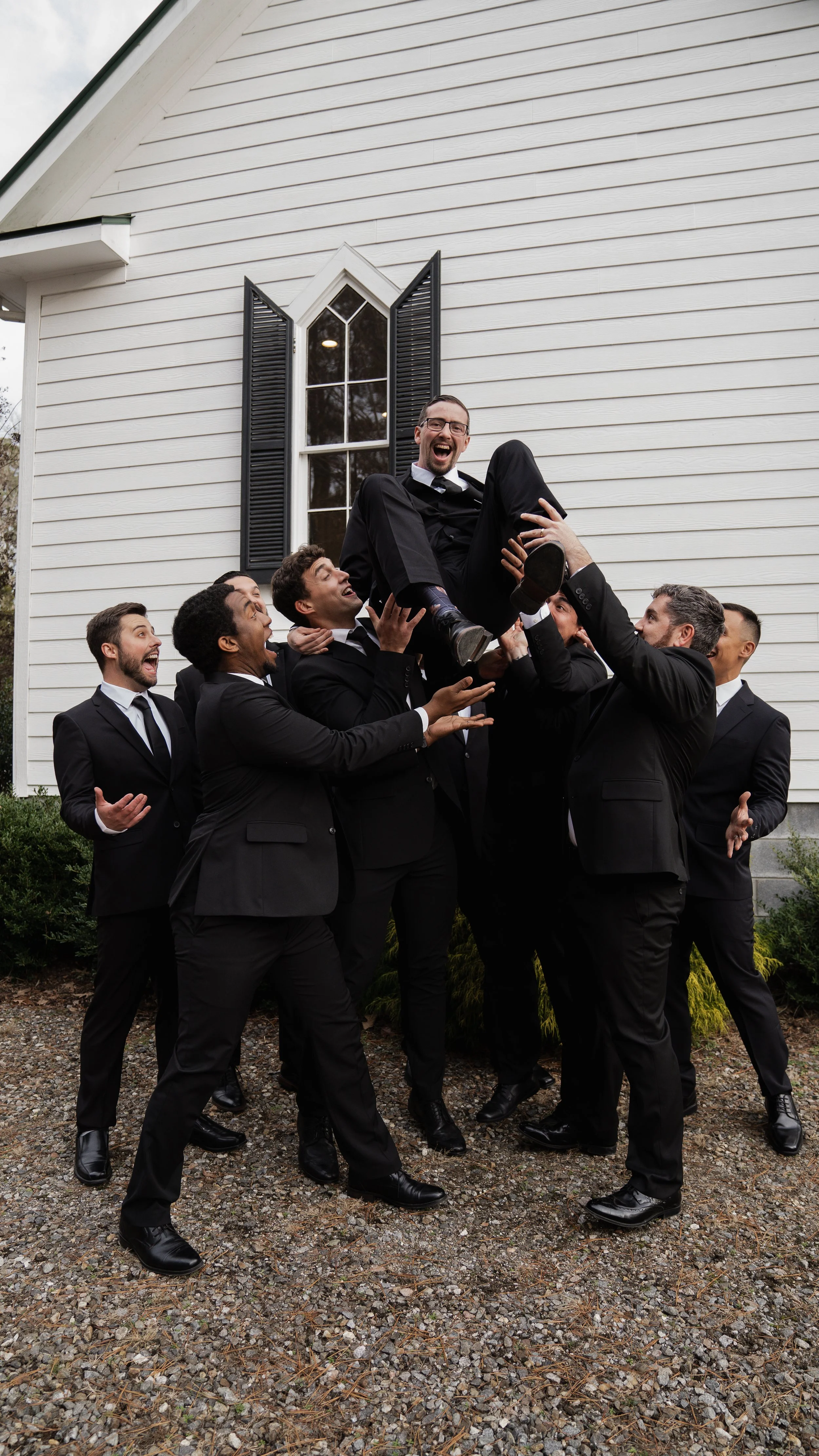 Group of men in black suits lifting a man in black suit and glasses, outdoors in front of white house with black shutters, celebrating.