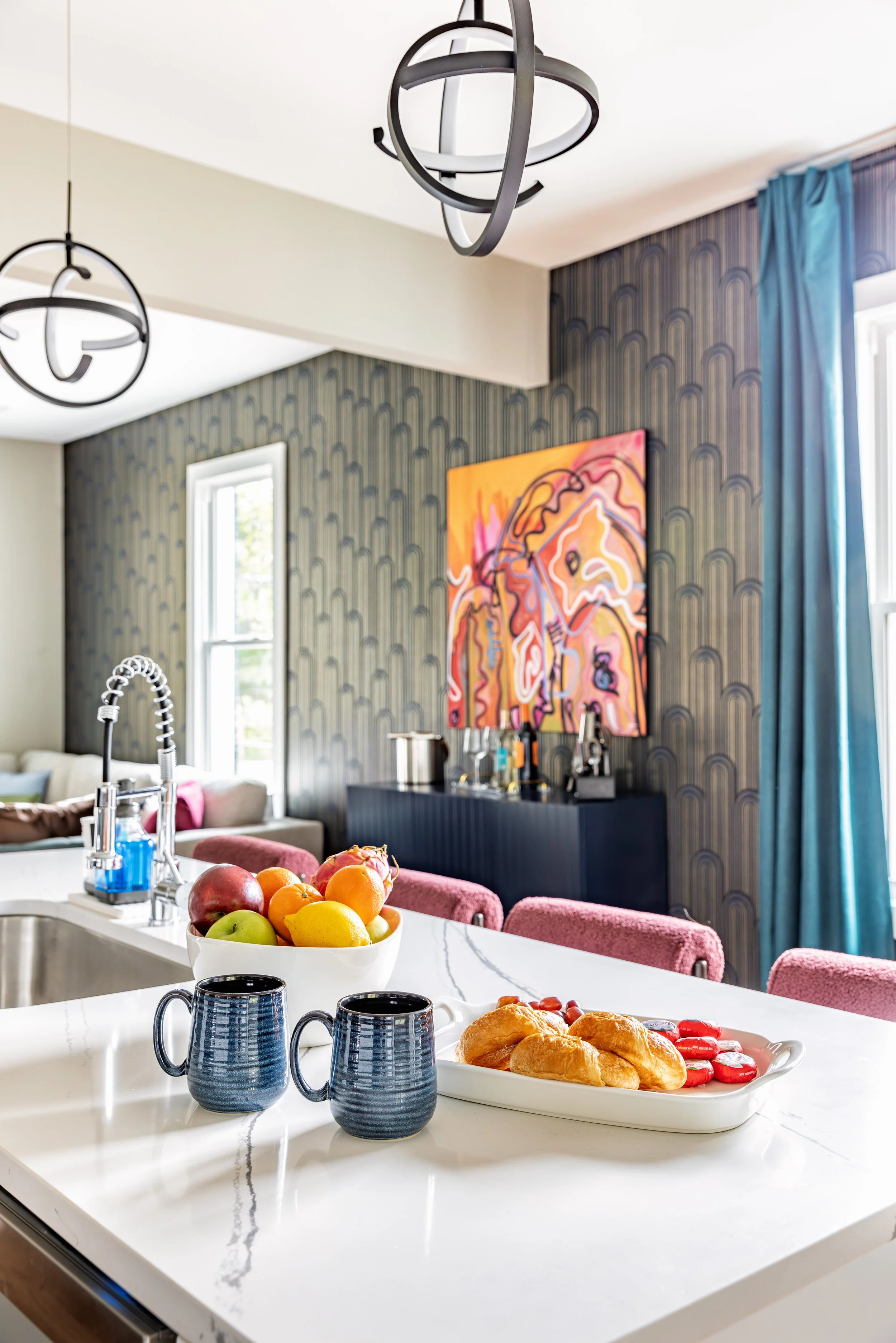 Modern kitchen and living area with breakfast spread, including a fruit bowl with apples, oranges, and a pomegranate, two blue mugs, and a tray of croissants and cookies on a white marble countertop. In the background, a colorful abstract painting and contemporary pendant lights are visible.