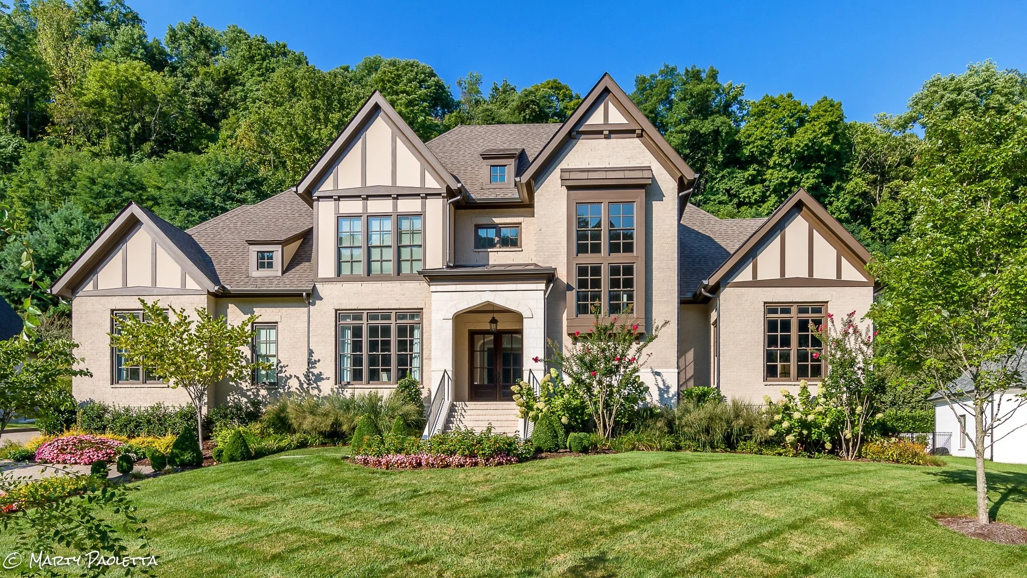 A large, modern house with a landscaped front yard, multiple roof gables, tall windows, and a pathway leading to the front door, surrounded by green trees and a bright blue sky.