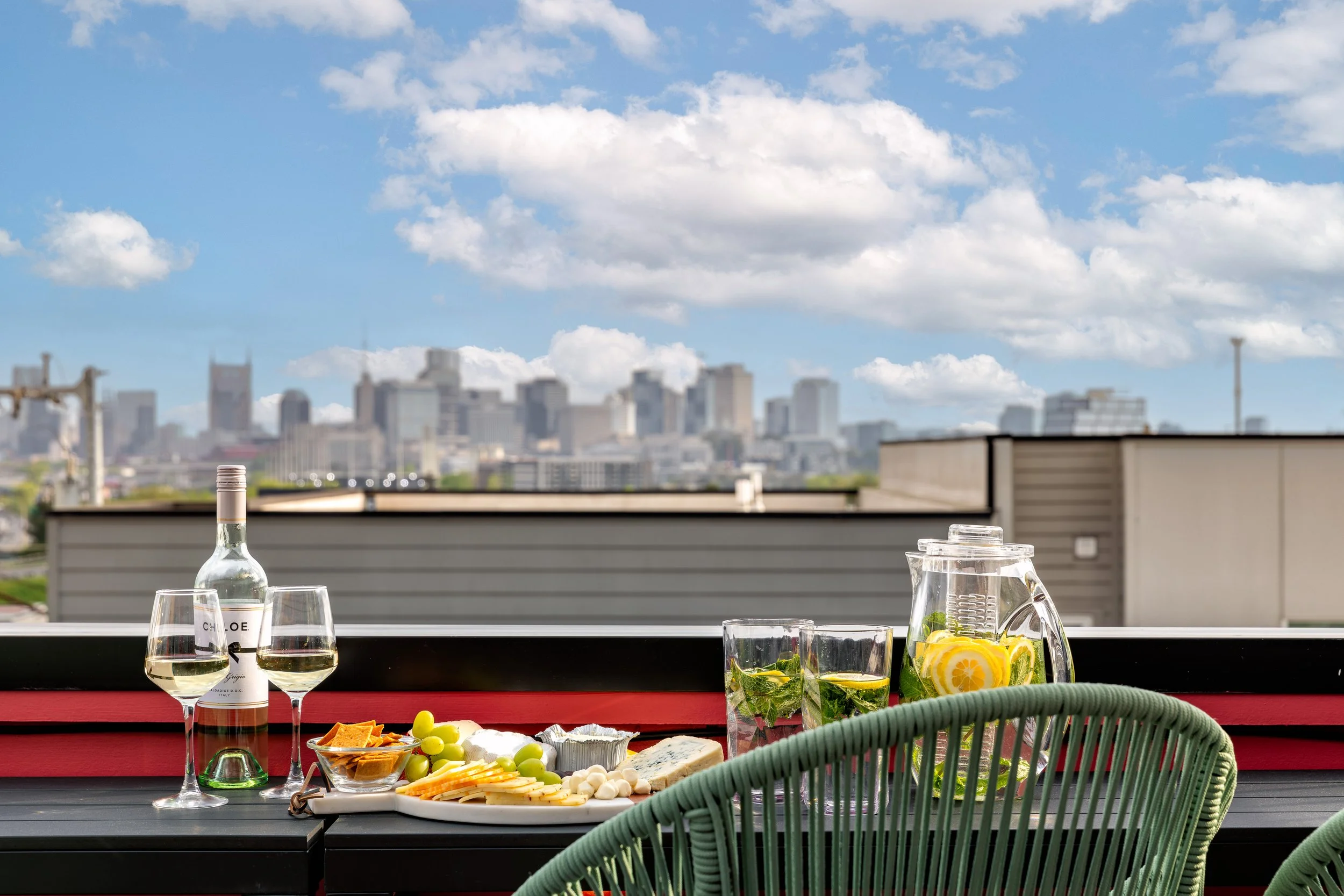Outdoor rooftop table set with wine, cheese, grapes, snacks, and lemonade, overlooking a city skyline under a partly cloudy sky.