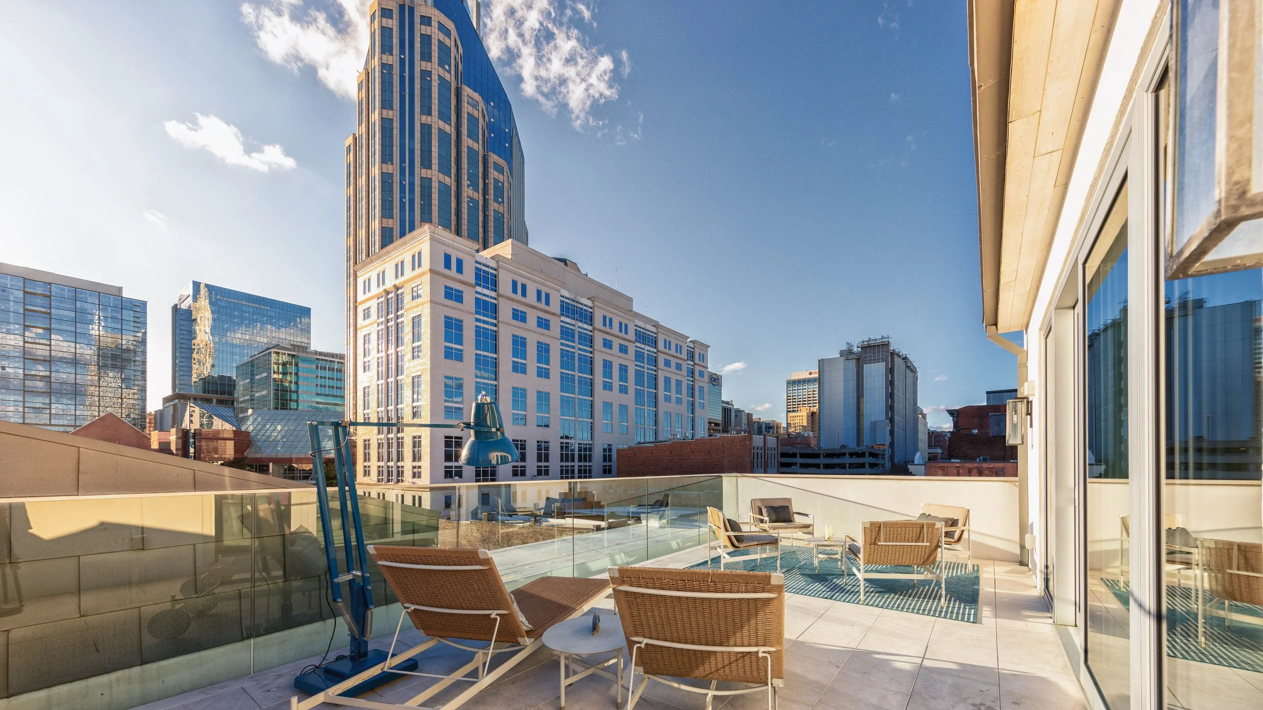 A modern rooftop patio in an urban cityscape with tables, chairs, and a glass railing, overlooking tall office buildings under a partly cloudy sky.