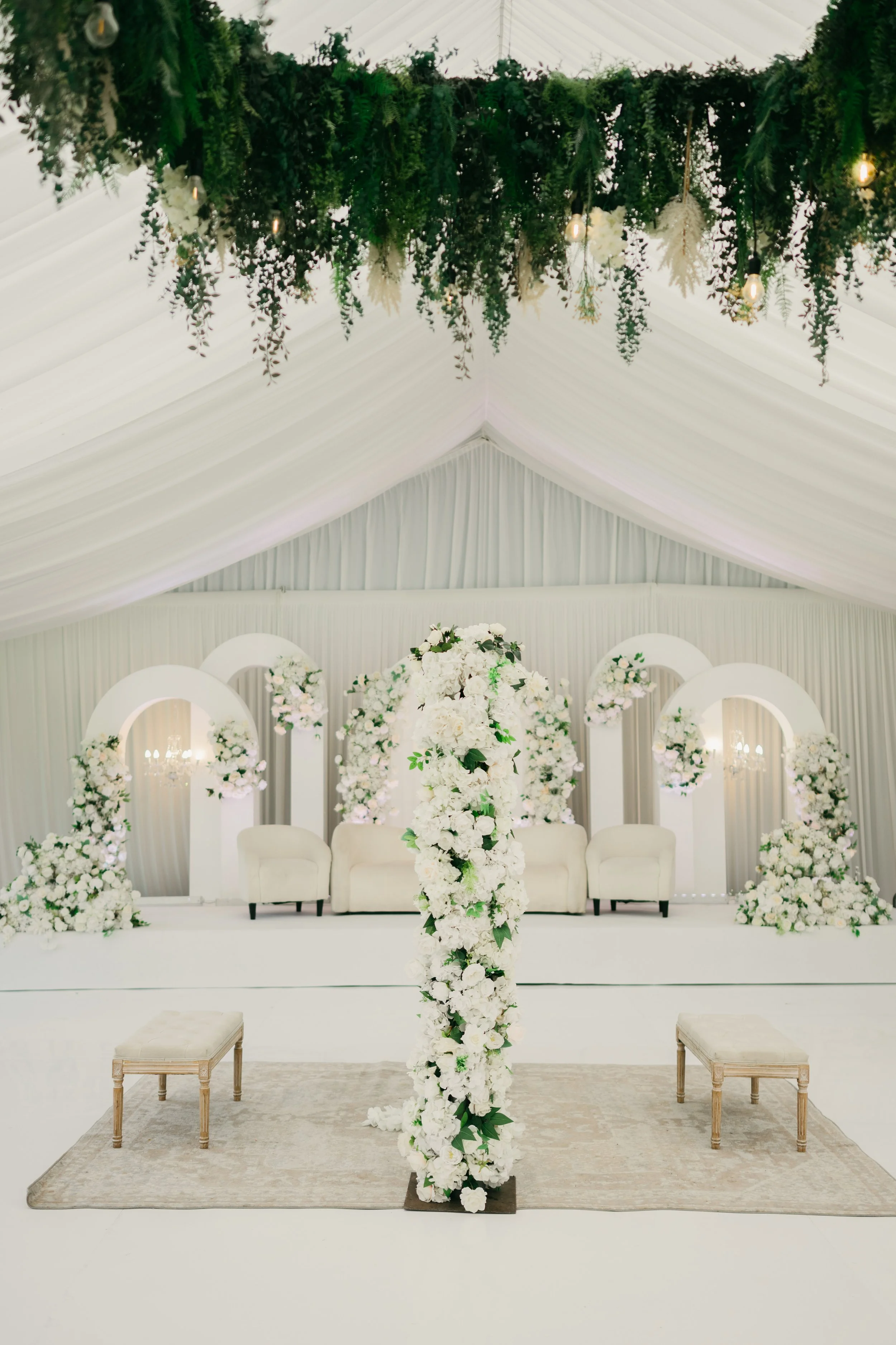A decorated wedding ceremony stage with white flowers, arches, and seating, under a white draped ceiling with hanging greenery and lights.