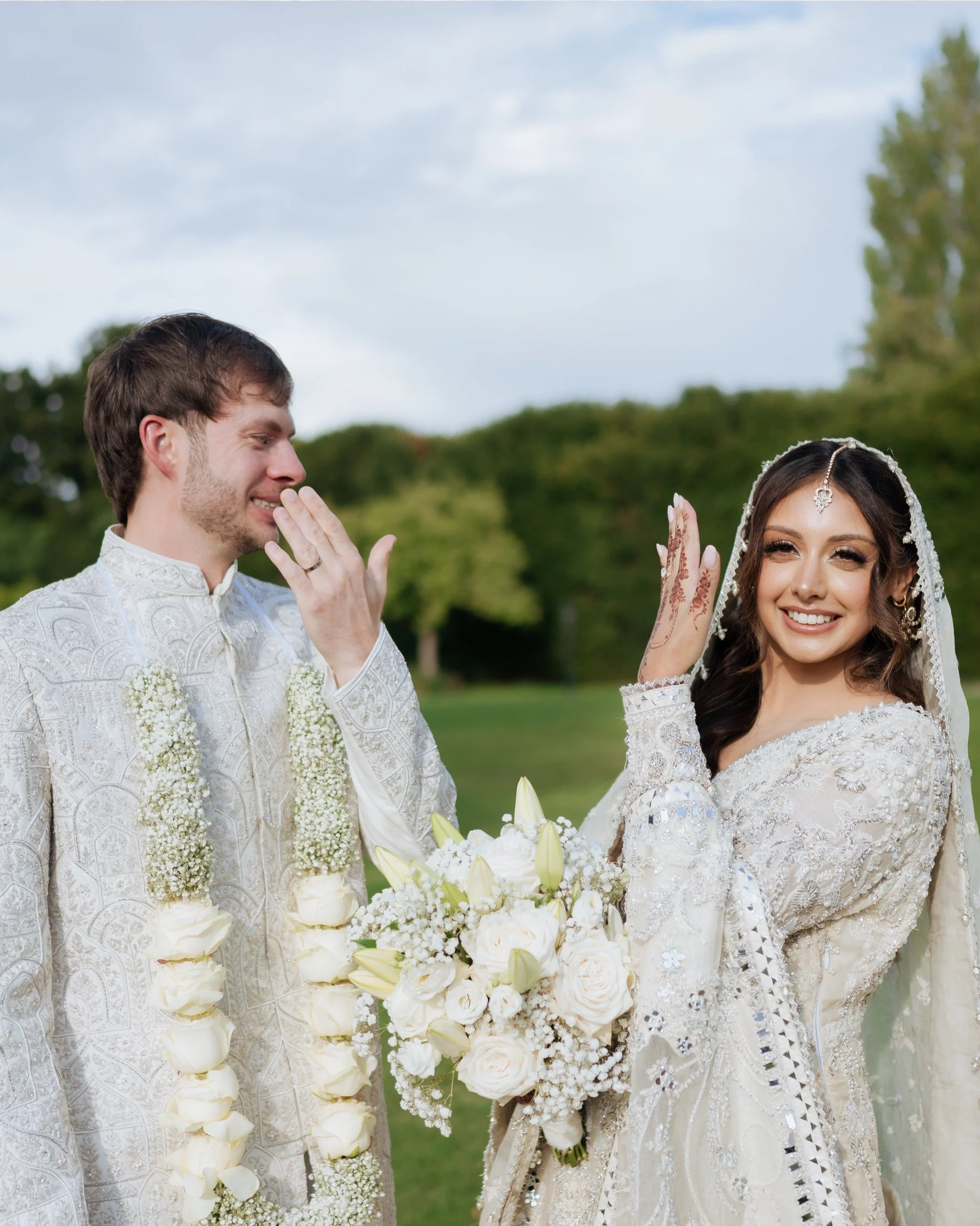Bride and groom in wedding attire smiling and showing their hands with a bouquet of white flowers in a lush outdoor setting.