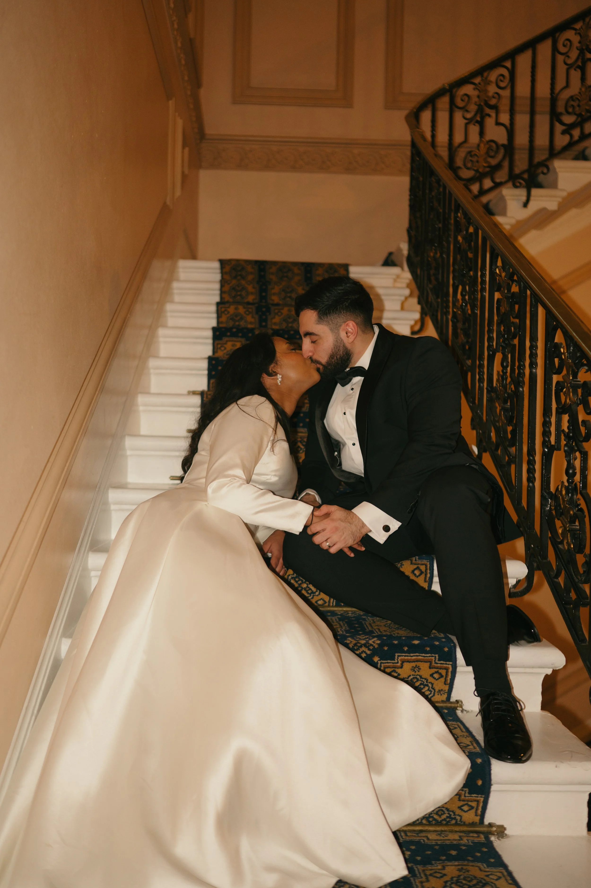 A bride and groom sharing a kiss on a staircase, with the bride in a white wedding gown and the groom in a black tuxedo, in an elegant indoor setting.
