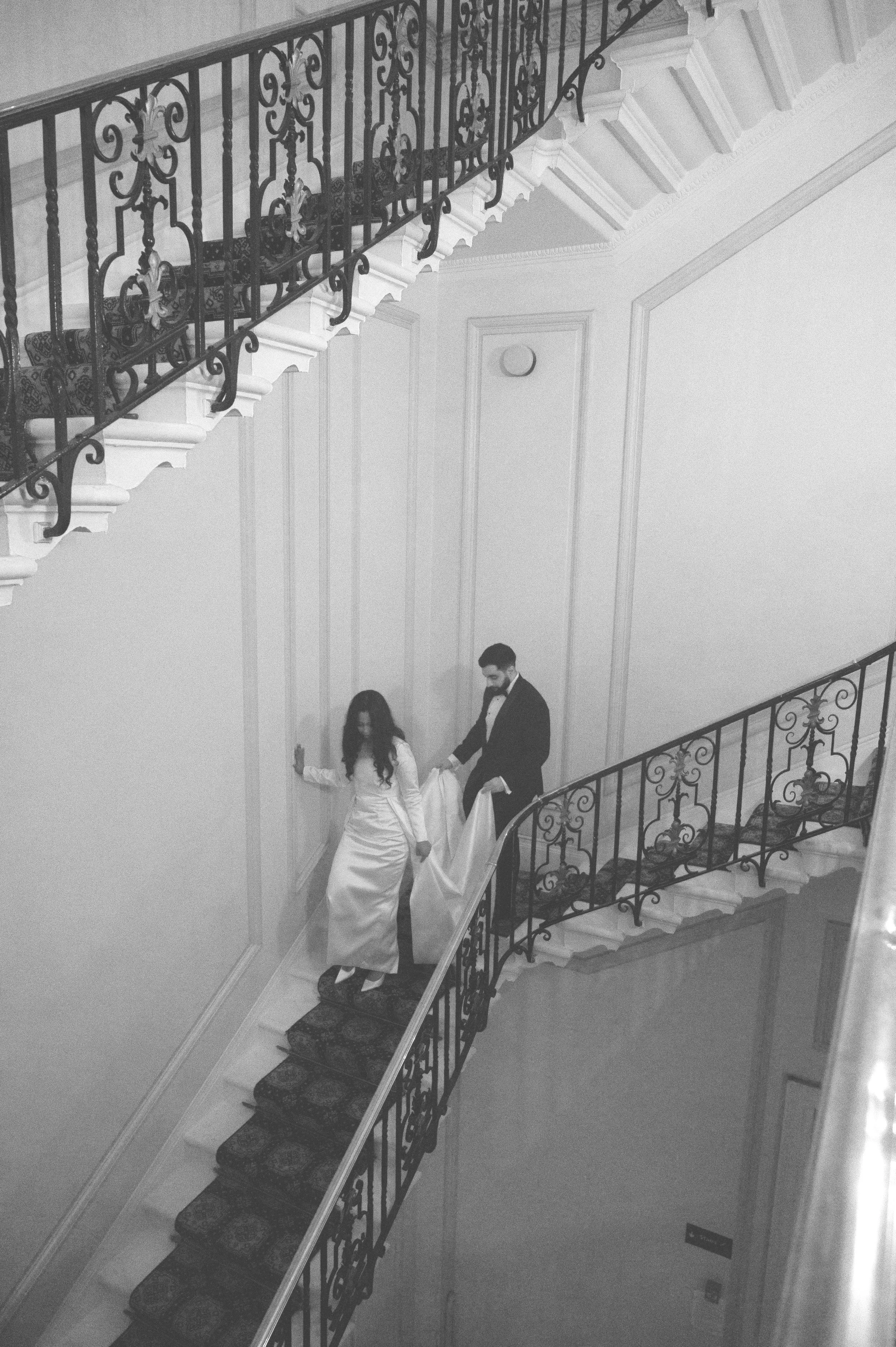 A bride and groom on a staircase, with the groom lifting the bride's dress as they walk down the stairs.