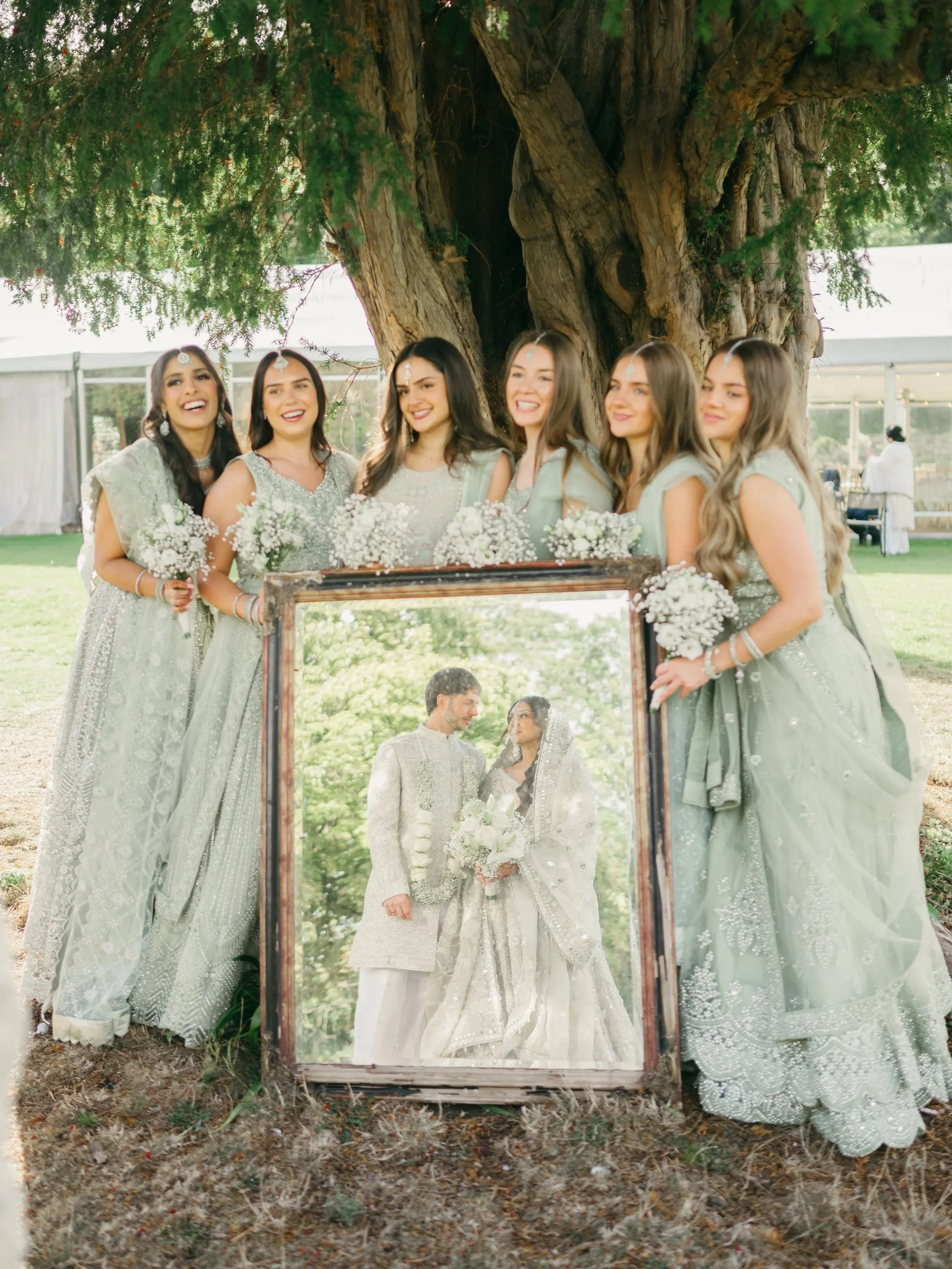 A group of bridesmaids in light green dresses standing outdoors under a large tree, holding bouquets of white flowers, with a mirror reflecting a bride and groom in wedding attire.