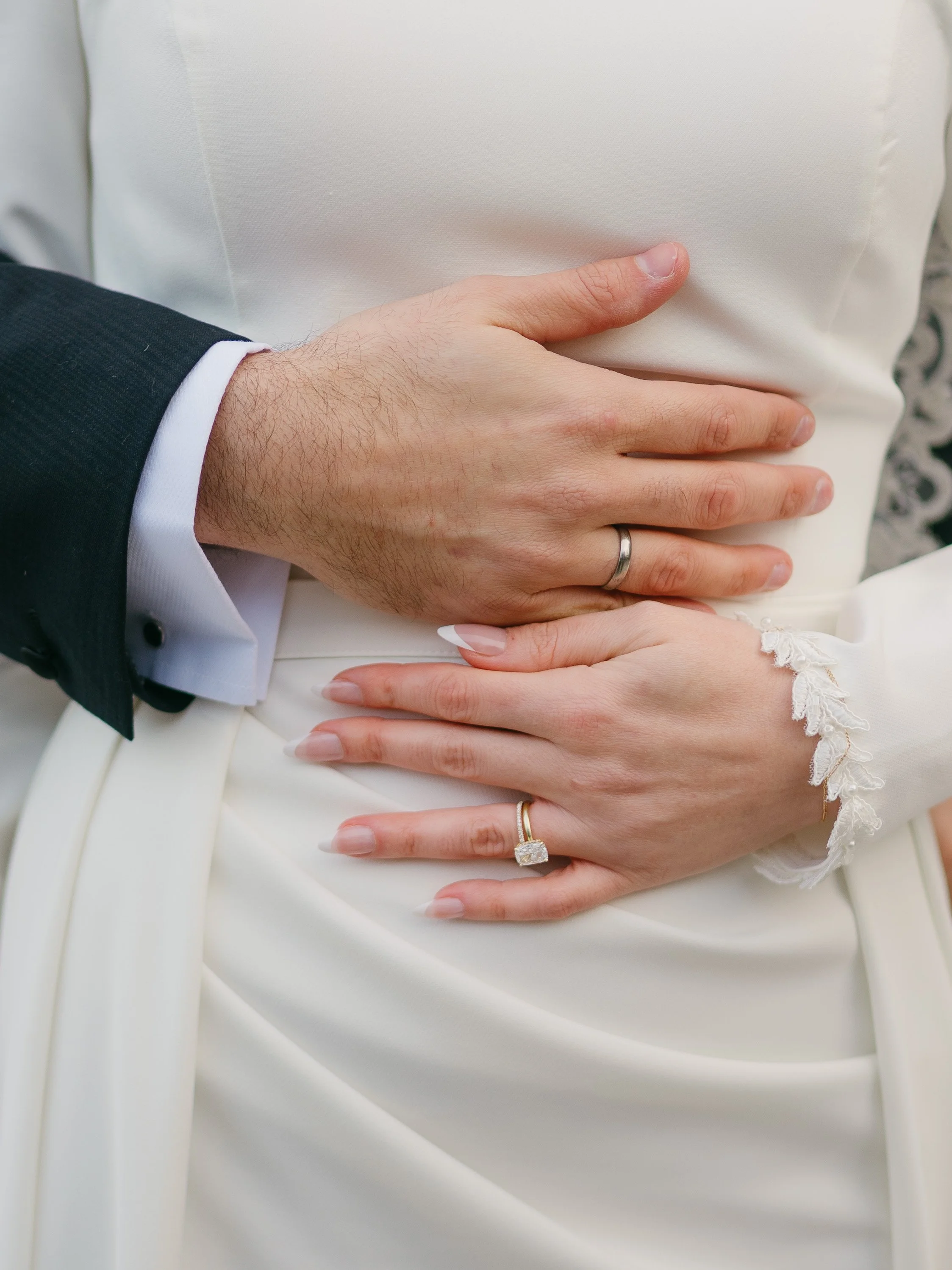Close-up of a married couple's hands resting on the bride's white wedding dress. The groom has a silver wedding band, and the bride has a gold engagement ring with a large diamond and a gold wedding band. The groom wears a dark suit jacket with a whi