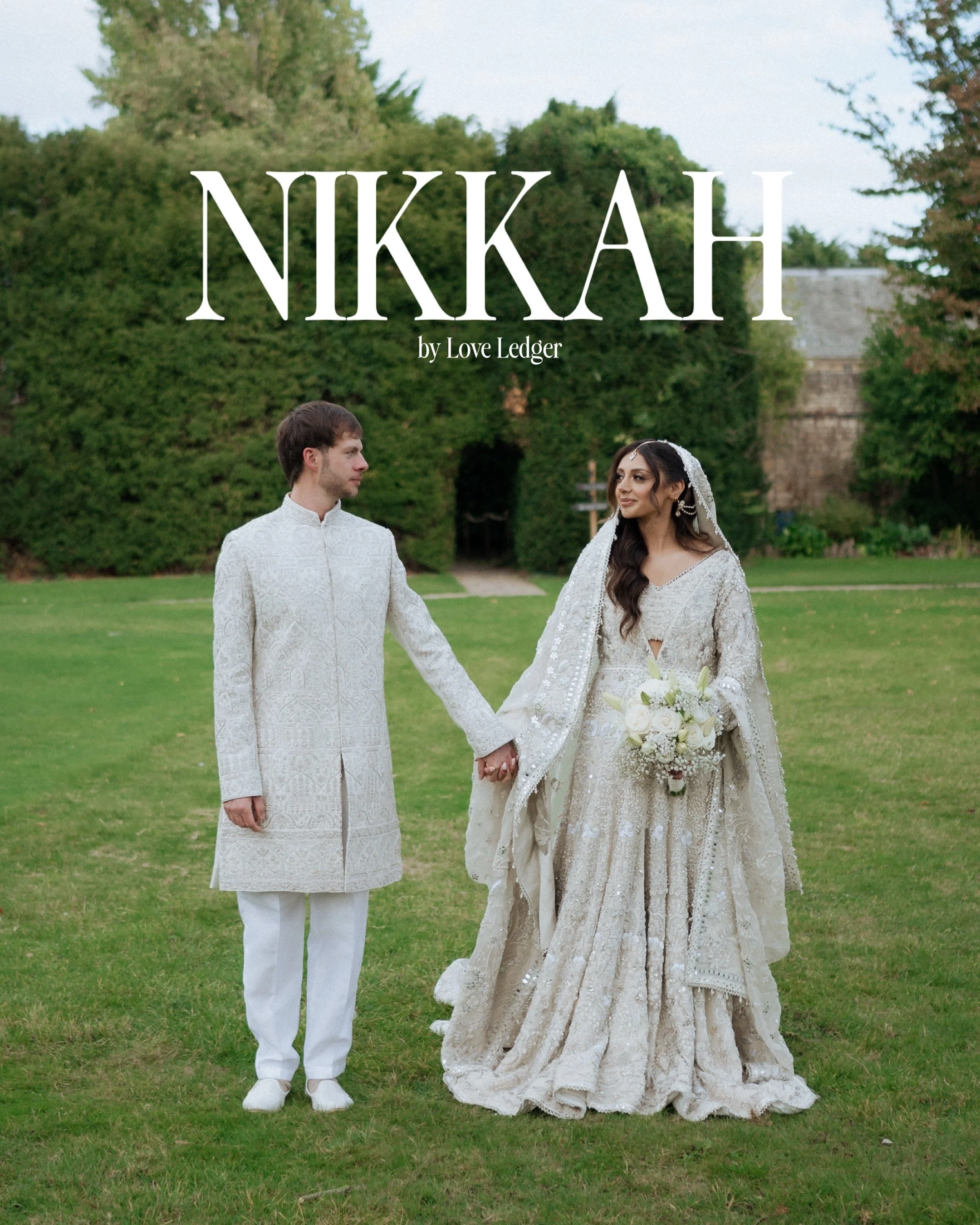 A couple dressed in wedding attire standing outdoors on a grassy field, holding hands, with trees and a building in the background.