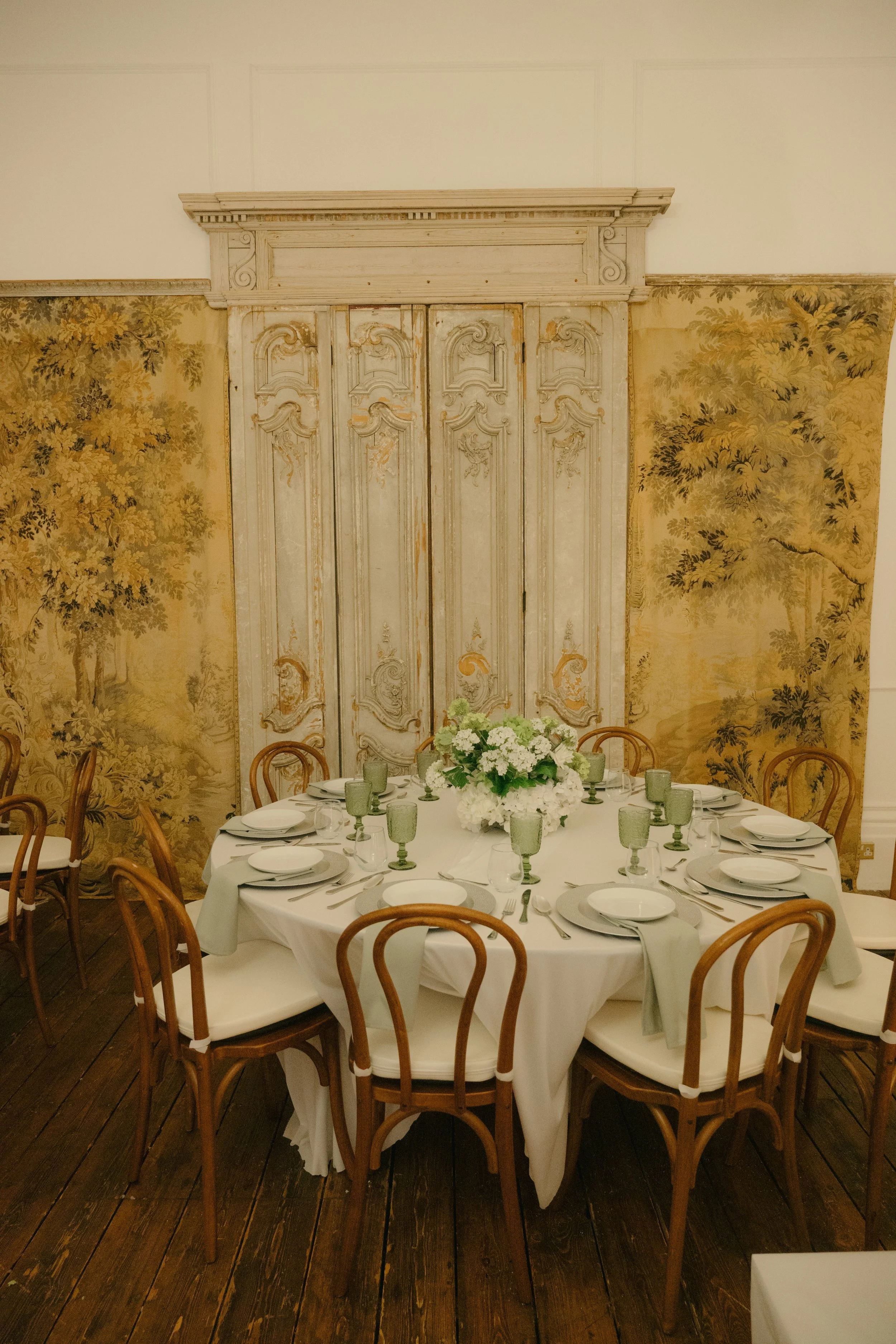A round dining table set for eight, with white tablecloth, white dinnerware, green goblets, and a floral centerpiece, in front of a vintage distressed door and ornate wallpaper depicting nature scenes.