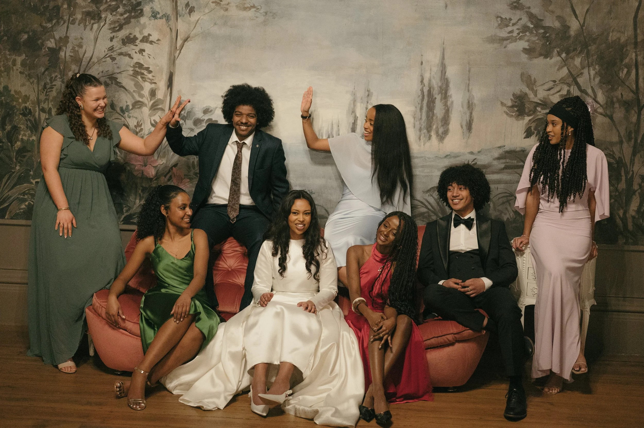 A group of nine people dressed in formal attire, sitting and standing in front of a painted backdrop of trees and a waterfall, smiling and celebrating.