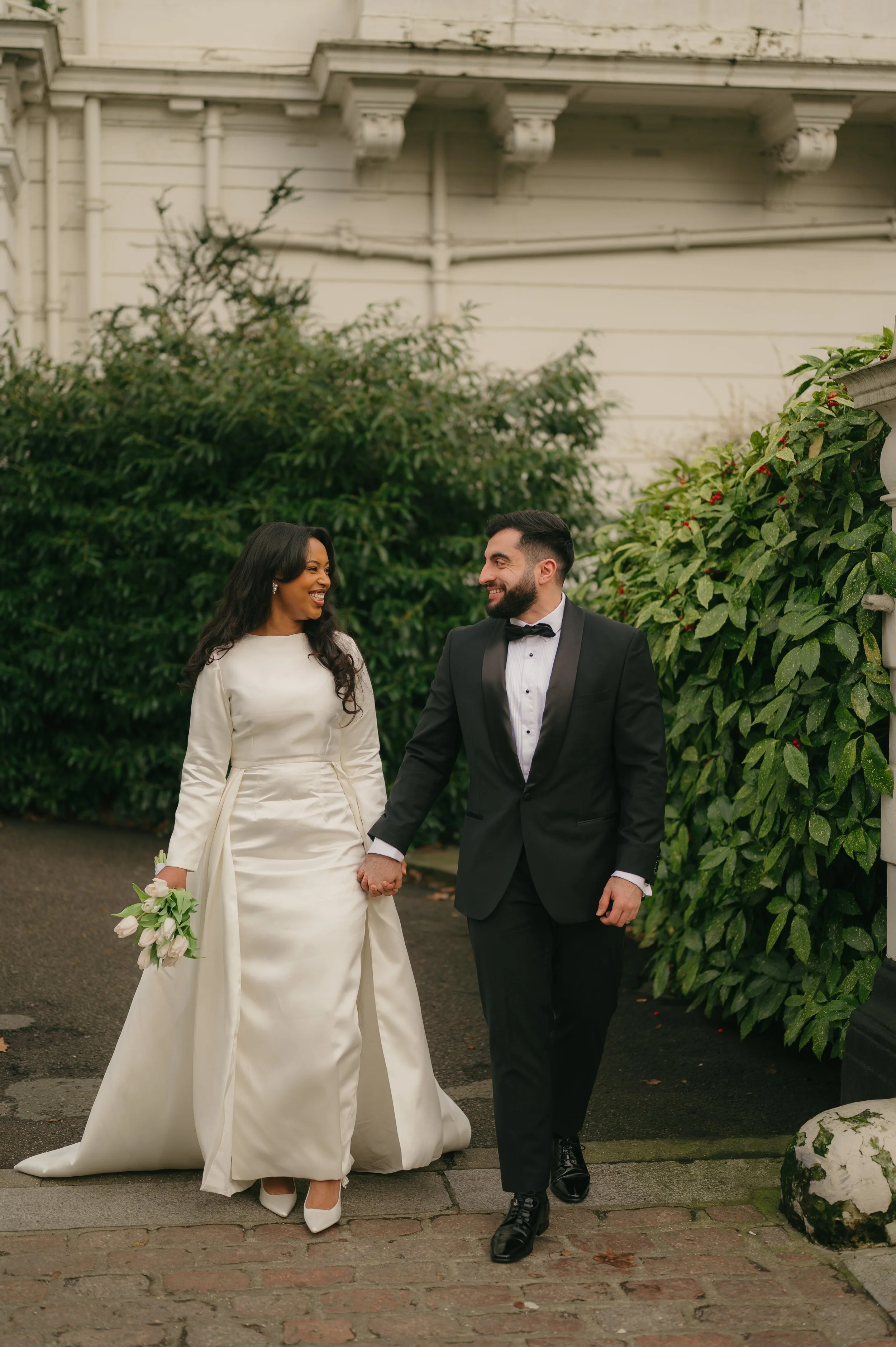 A newlywed couple holding hands and smiling at each other, walking outdoors on a brick pathway with lush greenery around them.