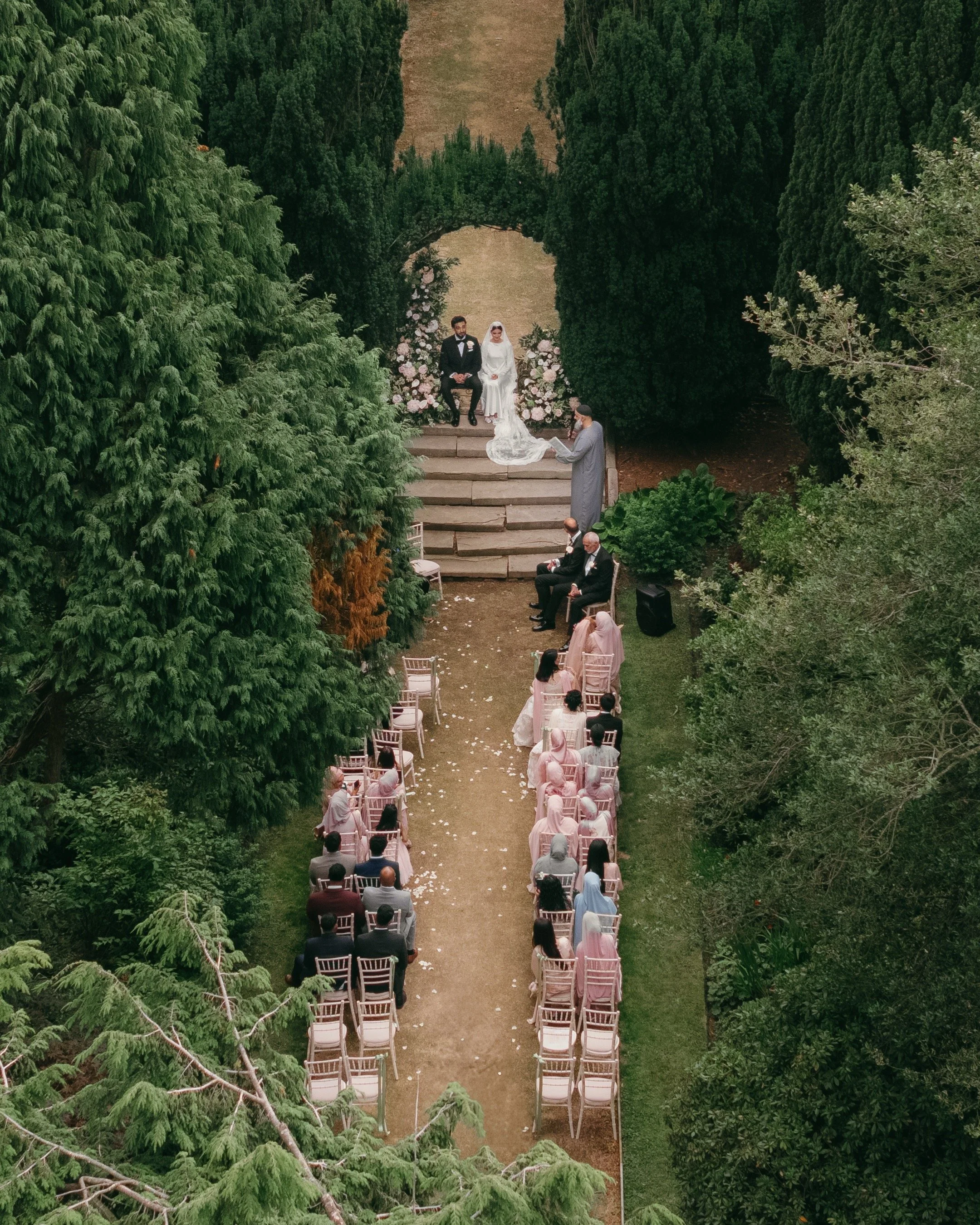 An outdoor wedding ceremony taking place among lush green trees, with a bride and groom seated on a decorated platform, officiant standing before them, and guests seated on chairs along an aisle lined with flower petals.
