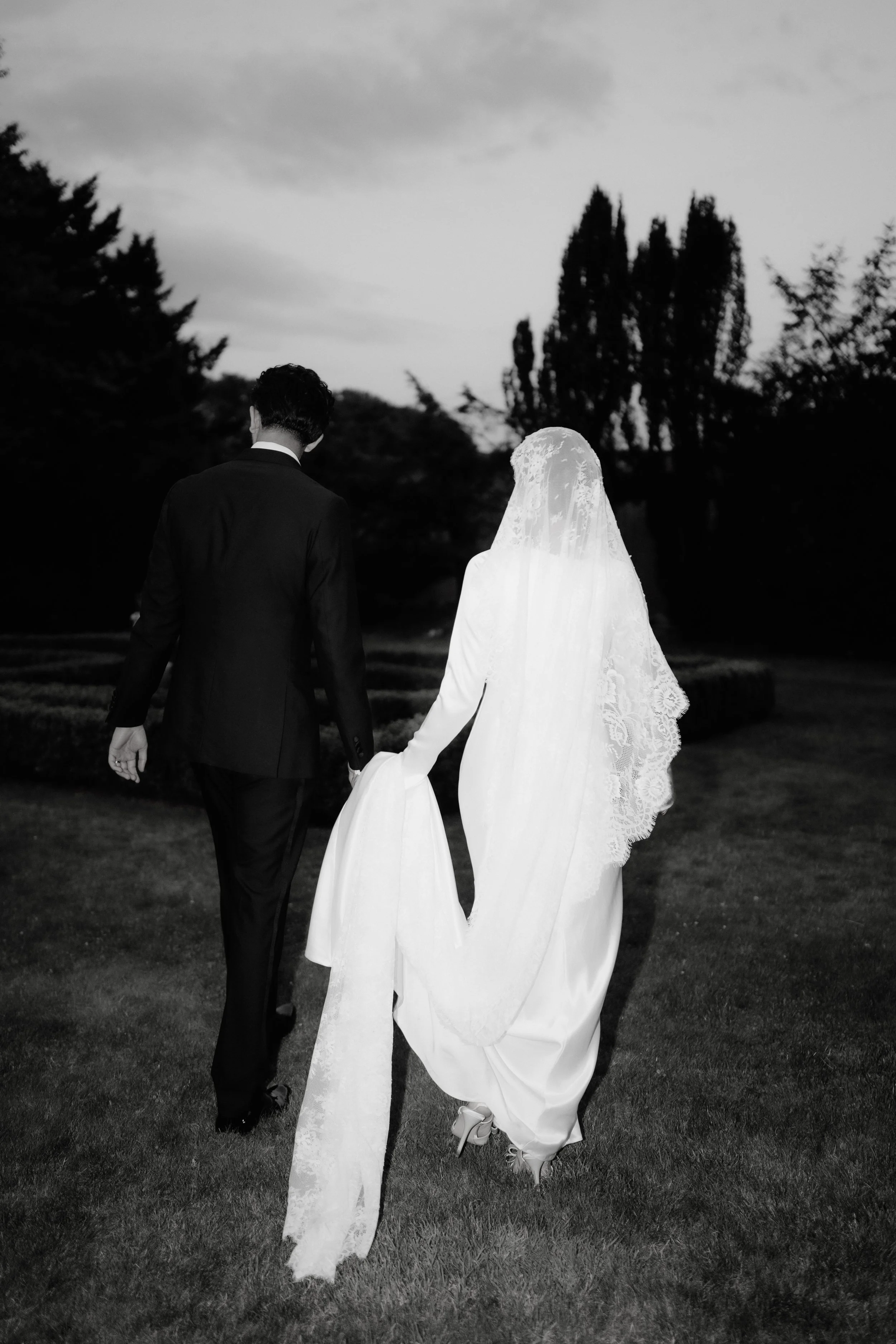 A bride and groom walking hand in hand across a grassy area, with trees in the background and the evening sky overhead.