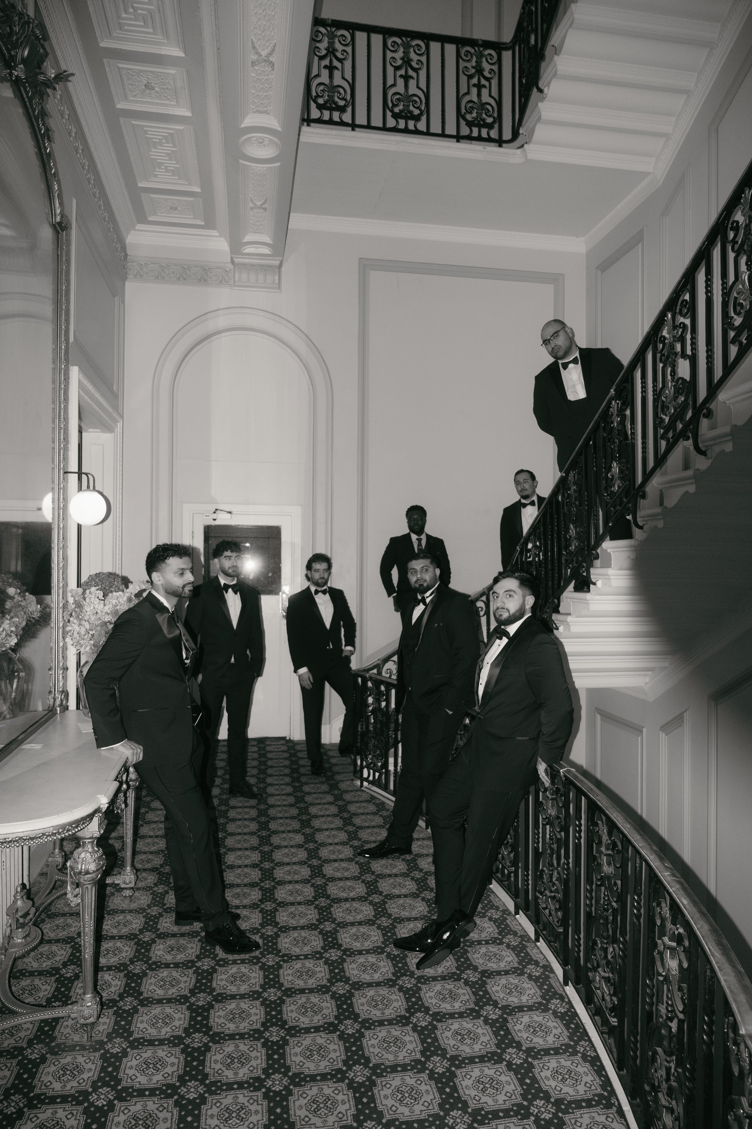 Group of seven men in tuxedos inside a fancy, historic building with ornate decor, floral arrangements, and patterned carpet, some standing on a staircase.