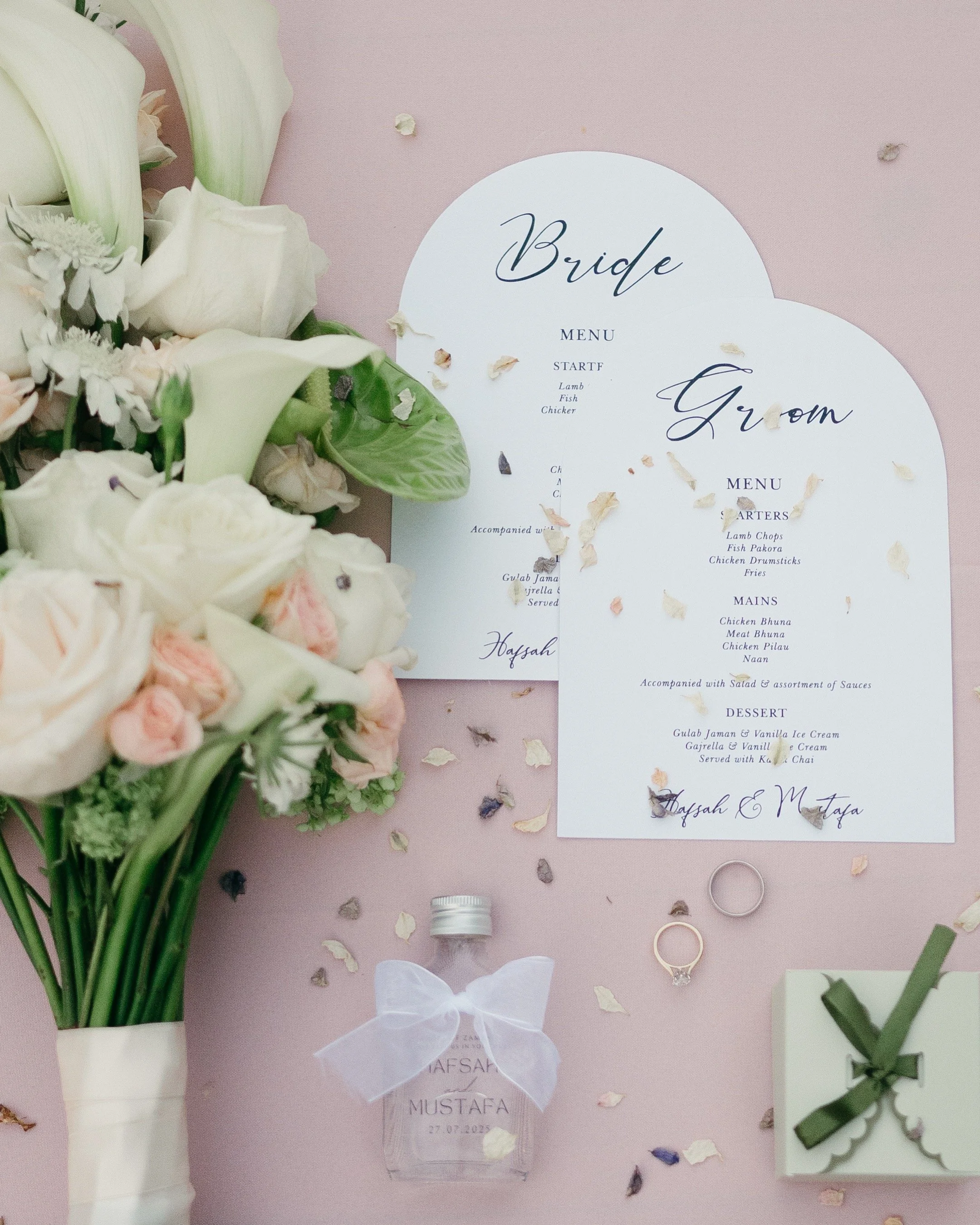 Wedding reception items on a pink table, including a bouquet of white and pink flowers, two heart-shaped wedding menus, two rings, a mini perfume bottle with a white bow, and a small wrapped gift box, with scattered flower petals.