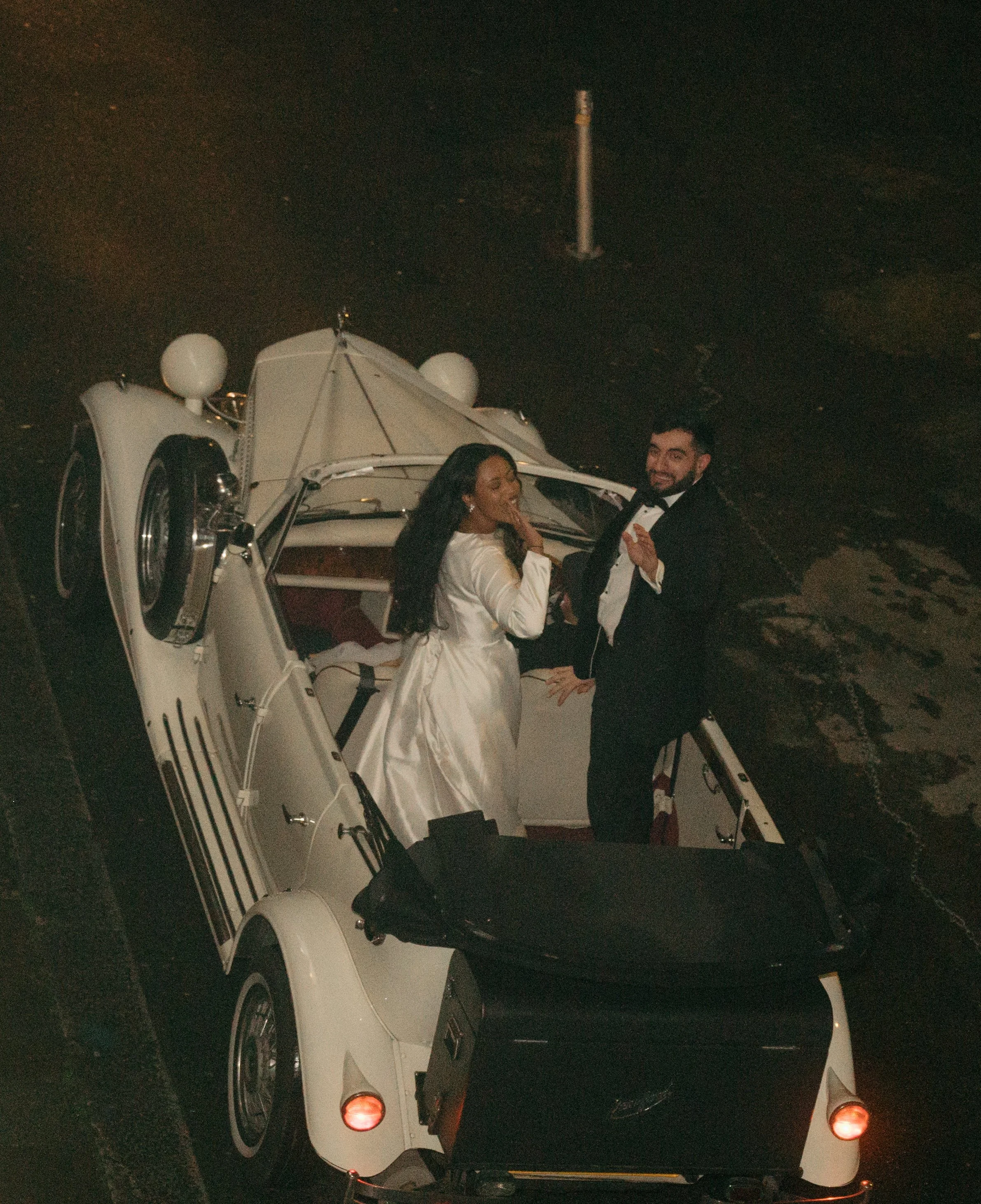 Couple dressed in tuxedo and wedding dress standing in golf cart on street at night.