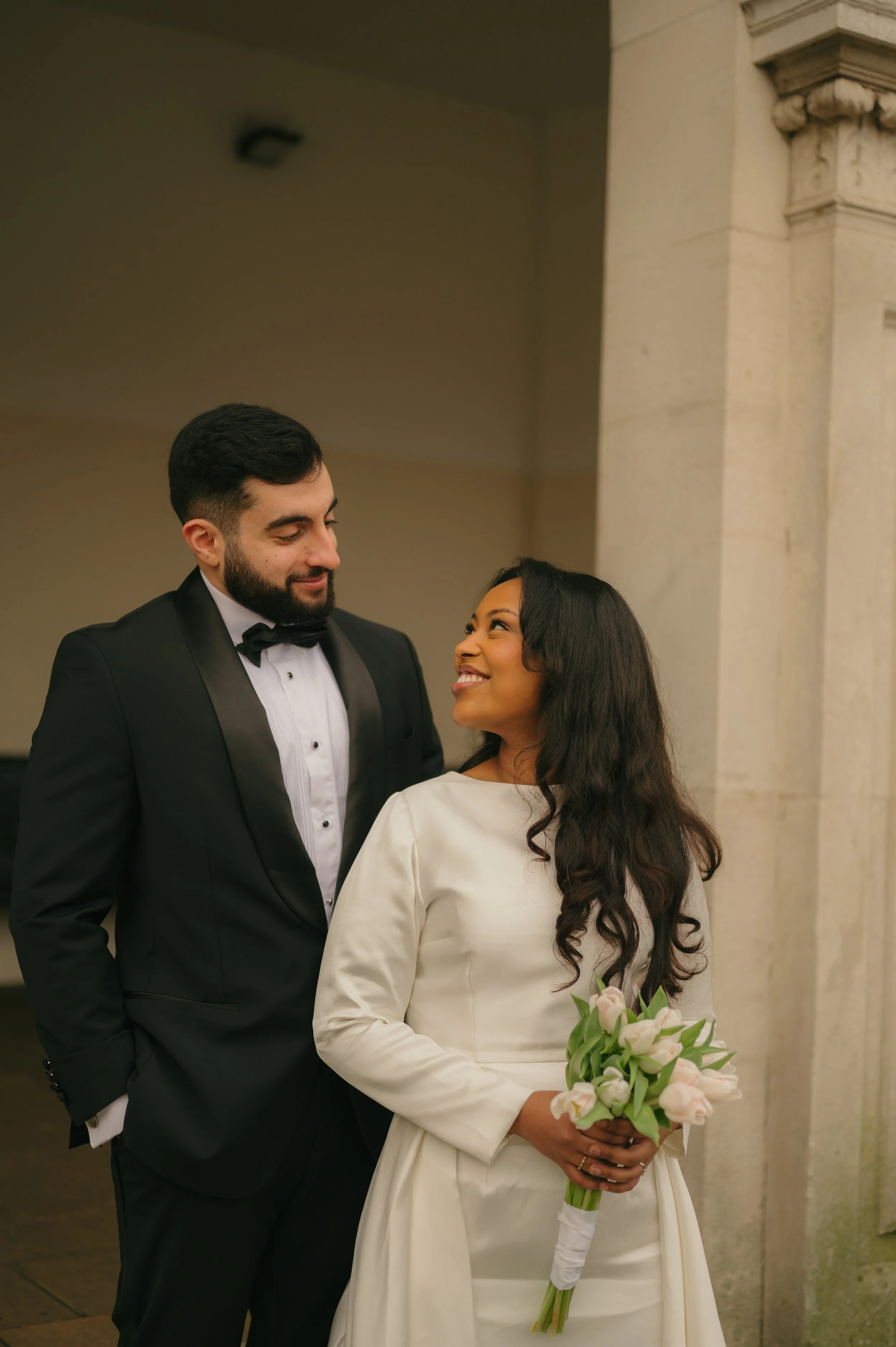 A bride and groom standing close together, smiling at each other, indoors. The bride wears a white wedding dress and holds a bouquet of pink and white tulips. The groom wears a black tuxedo with a bow tie.