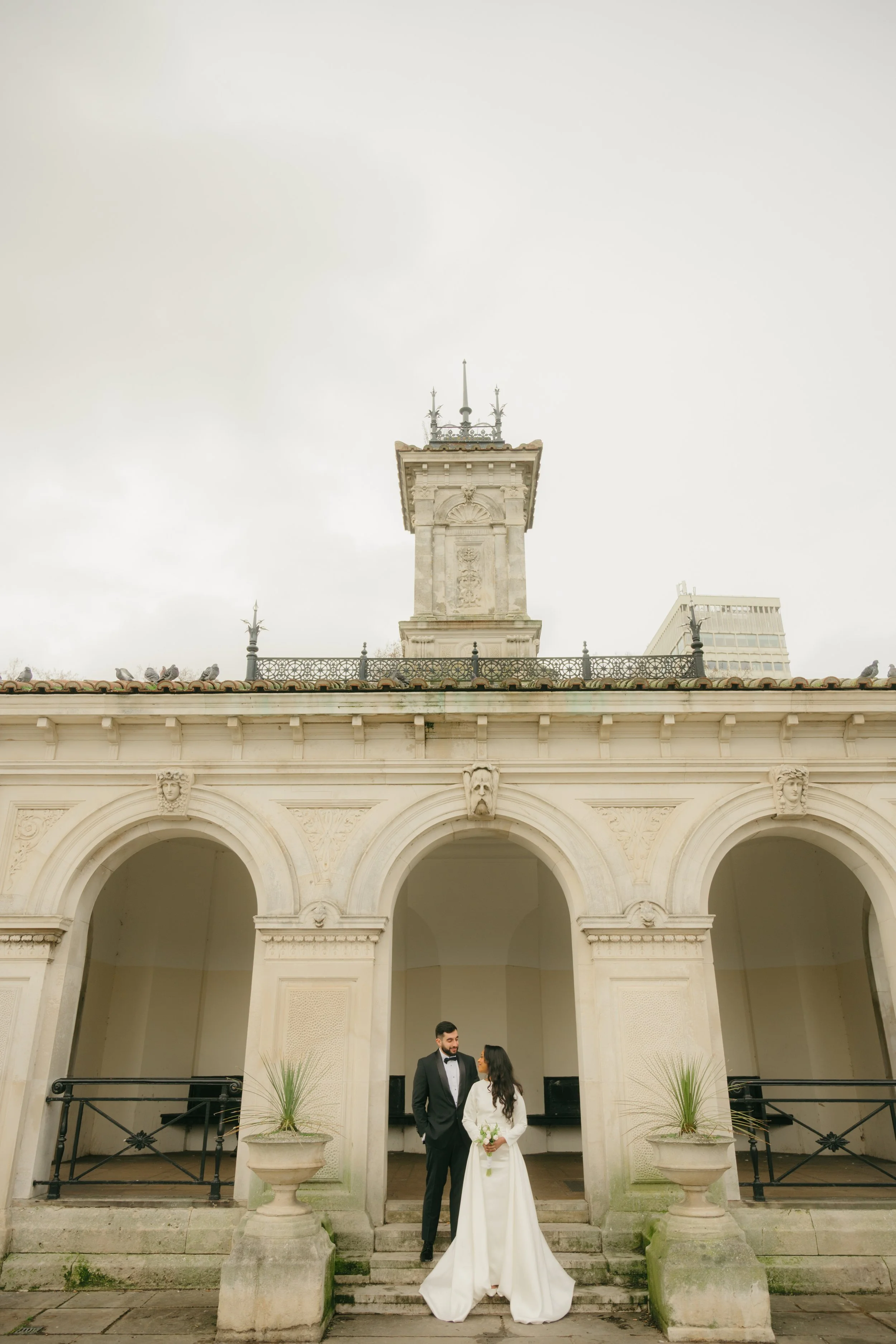 A bride and groom standing on steps outside a historic building with arched entrances and decorative stonework, overcast sky in the background.