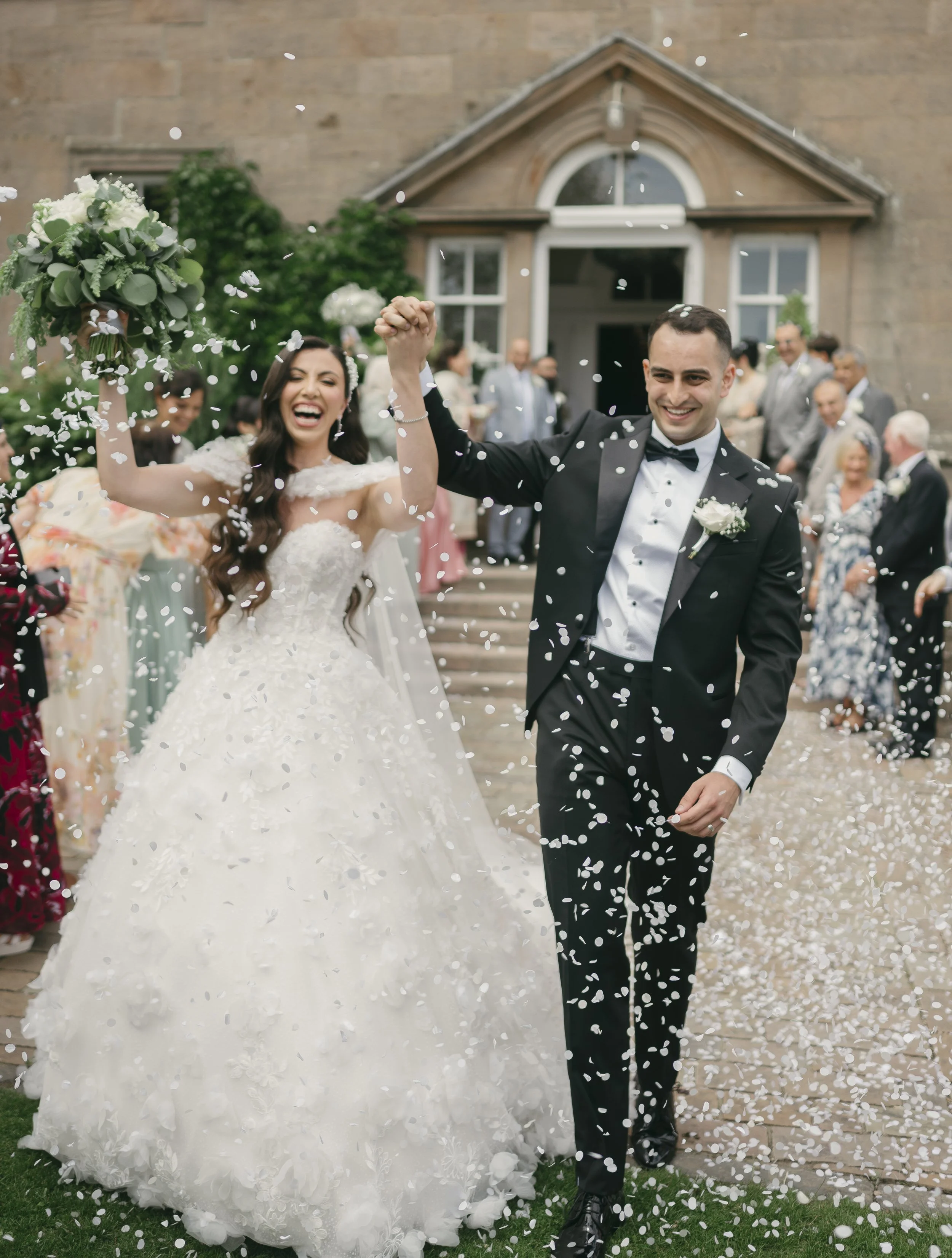 Wedding couple, bride in wedding gown and groom in tuxedo, celebrating with guests outside church with confetti.