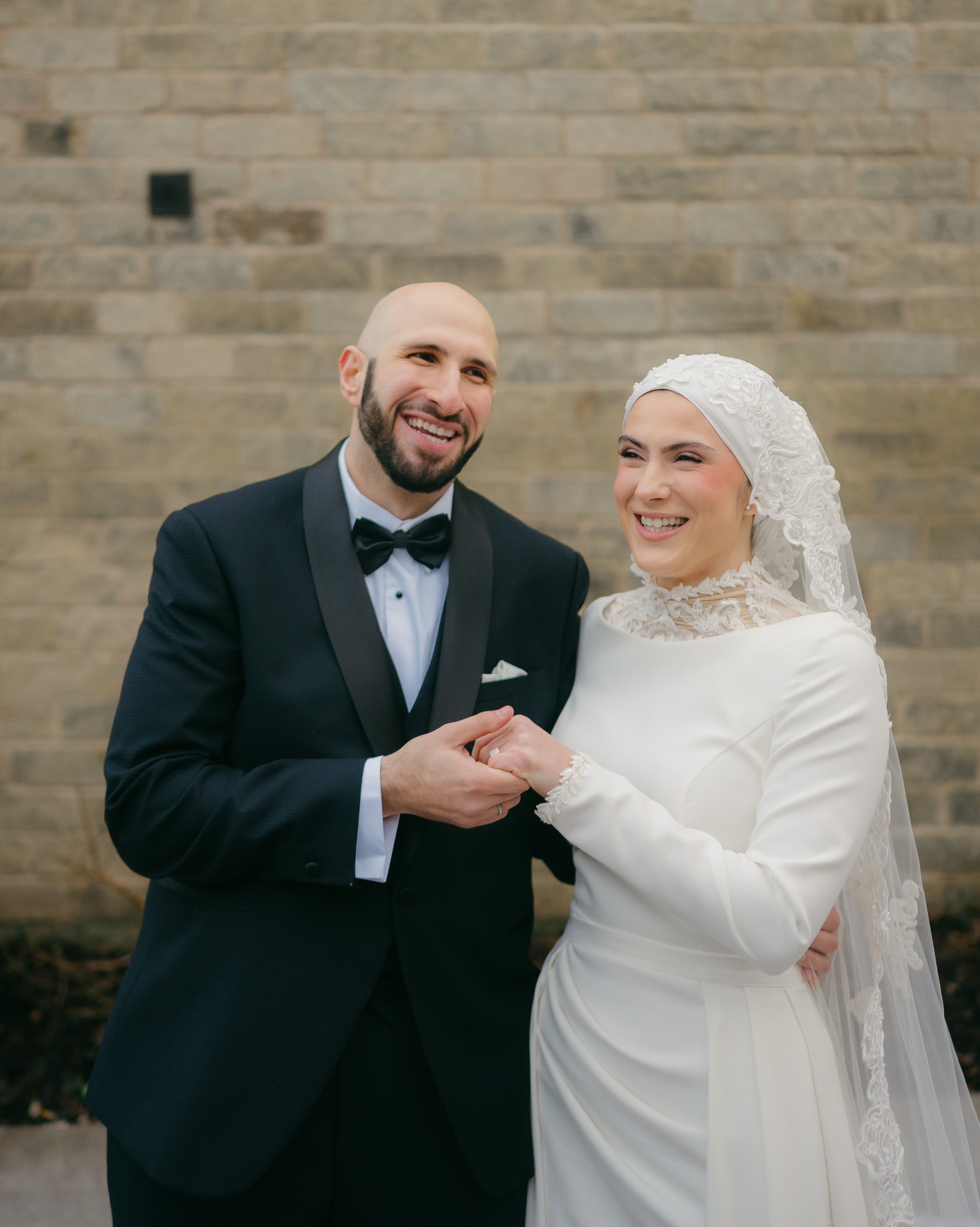 A newlywed couple smiling and holding hands in front of a brick wall, with the groom wearing a black tuxedo and the bride wearing a white wedding dress with lace details and a lace head covering.