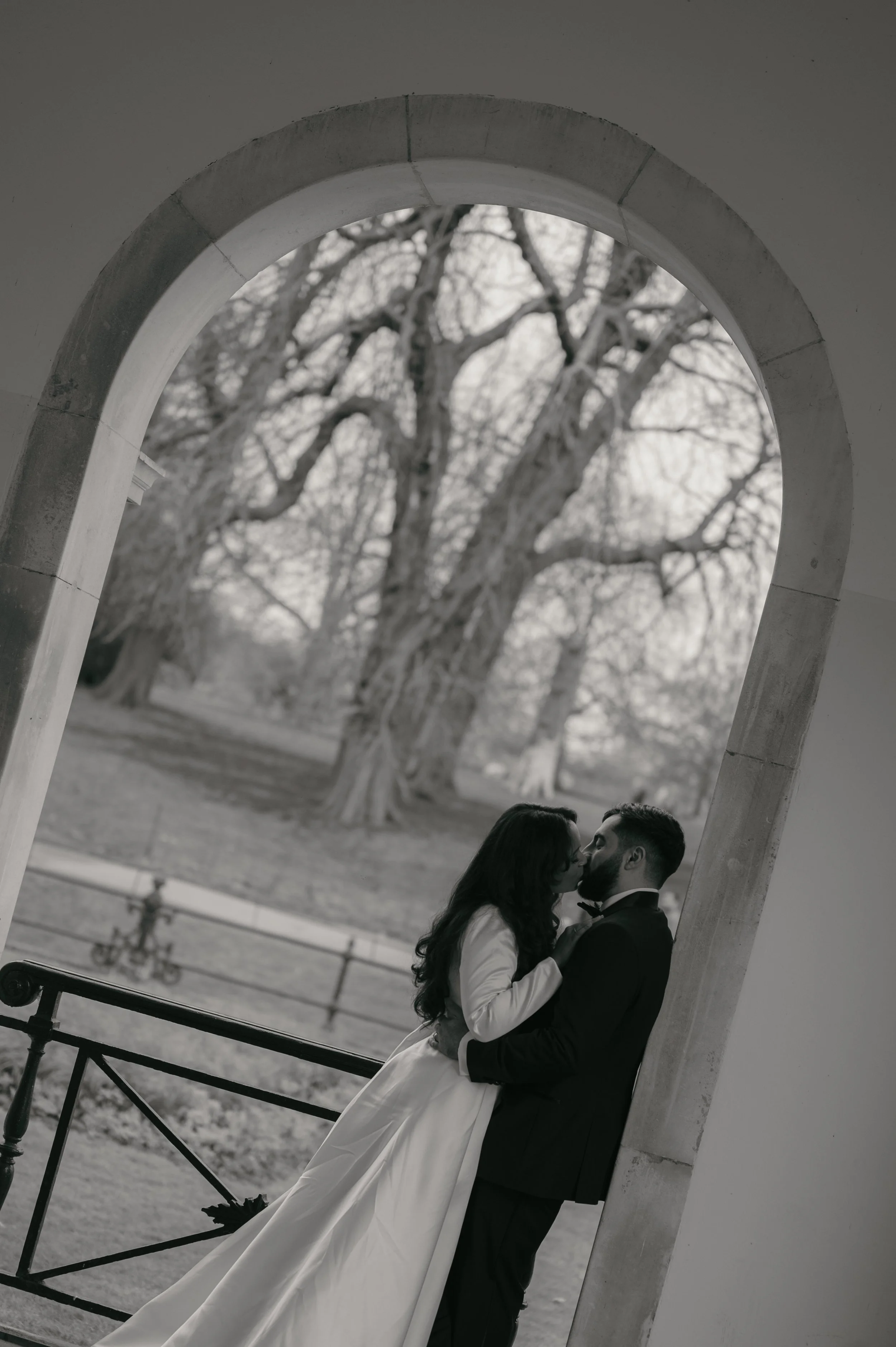 A couple dressed in wedding attire kissing under an arched opening with trees in the background.