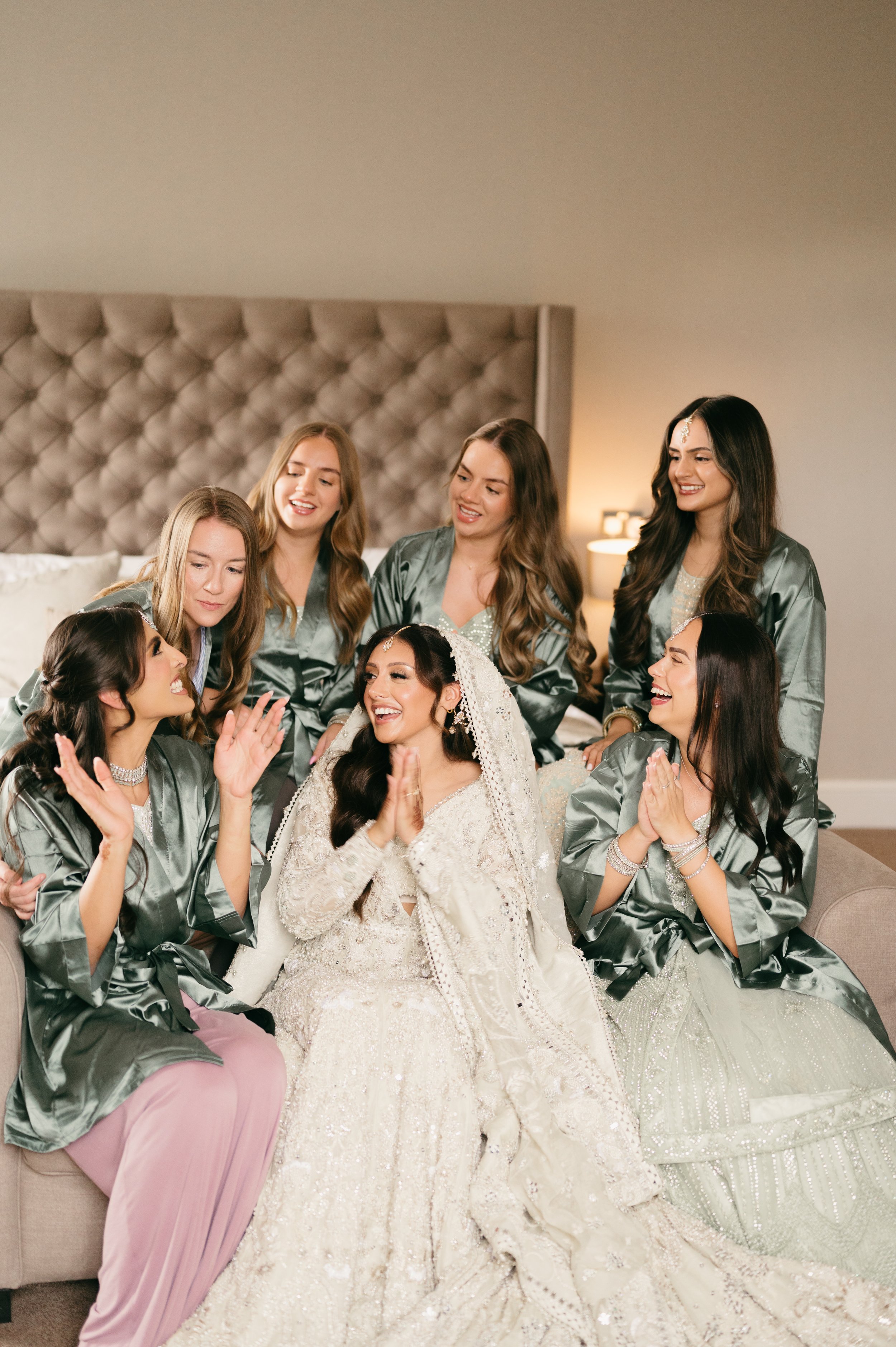 Bride and six bridesmaids in matching satin robes laughing and celebrating in a hotel room, with the bride in a wedding dress and veil.