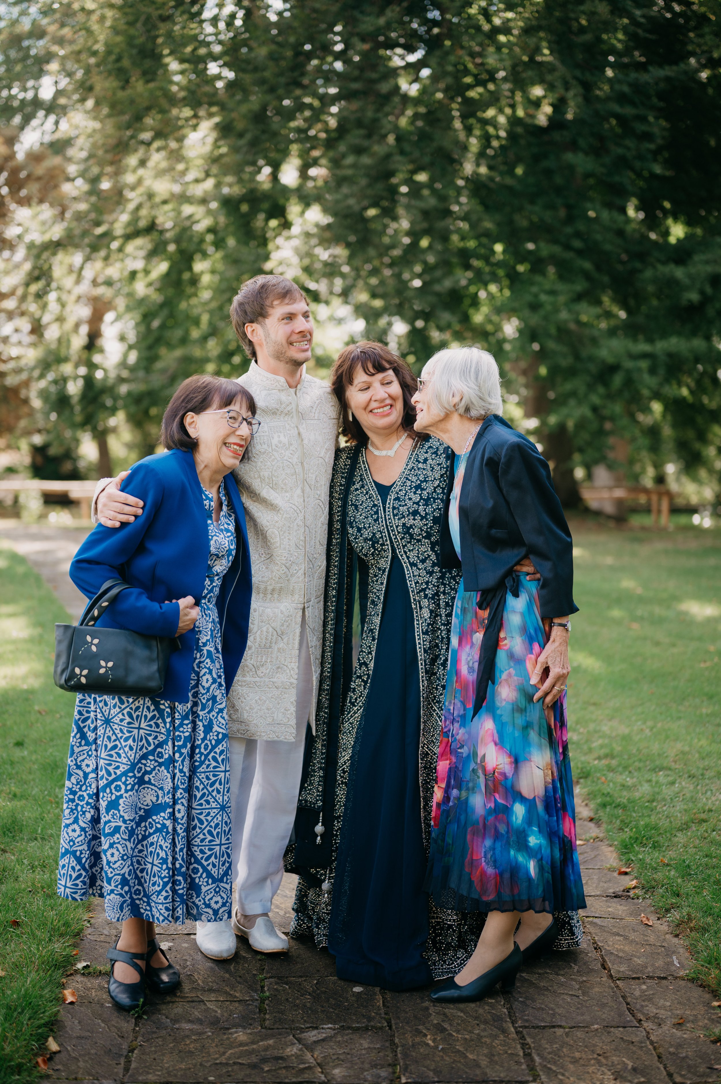 Group of five smiling people, two middle-aged women, a young man, and an elderly woman sharing a hug outdoors on a grassy area, with large trees and dappled sunlight in the background.