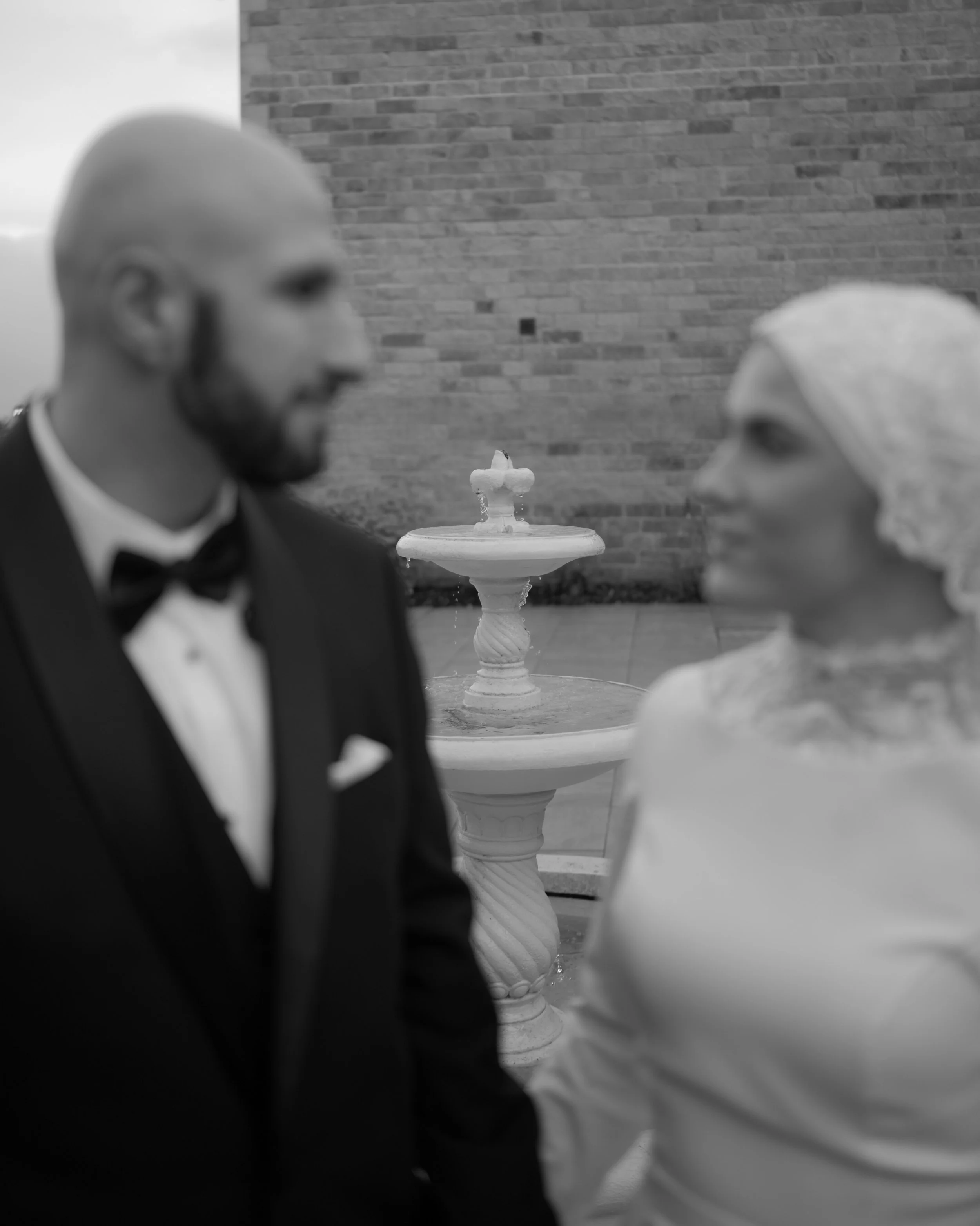 A black and white photo of a tuxedoed man and woman facing each other outdoors with a fountain in the background.