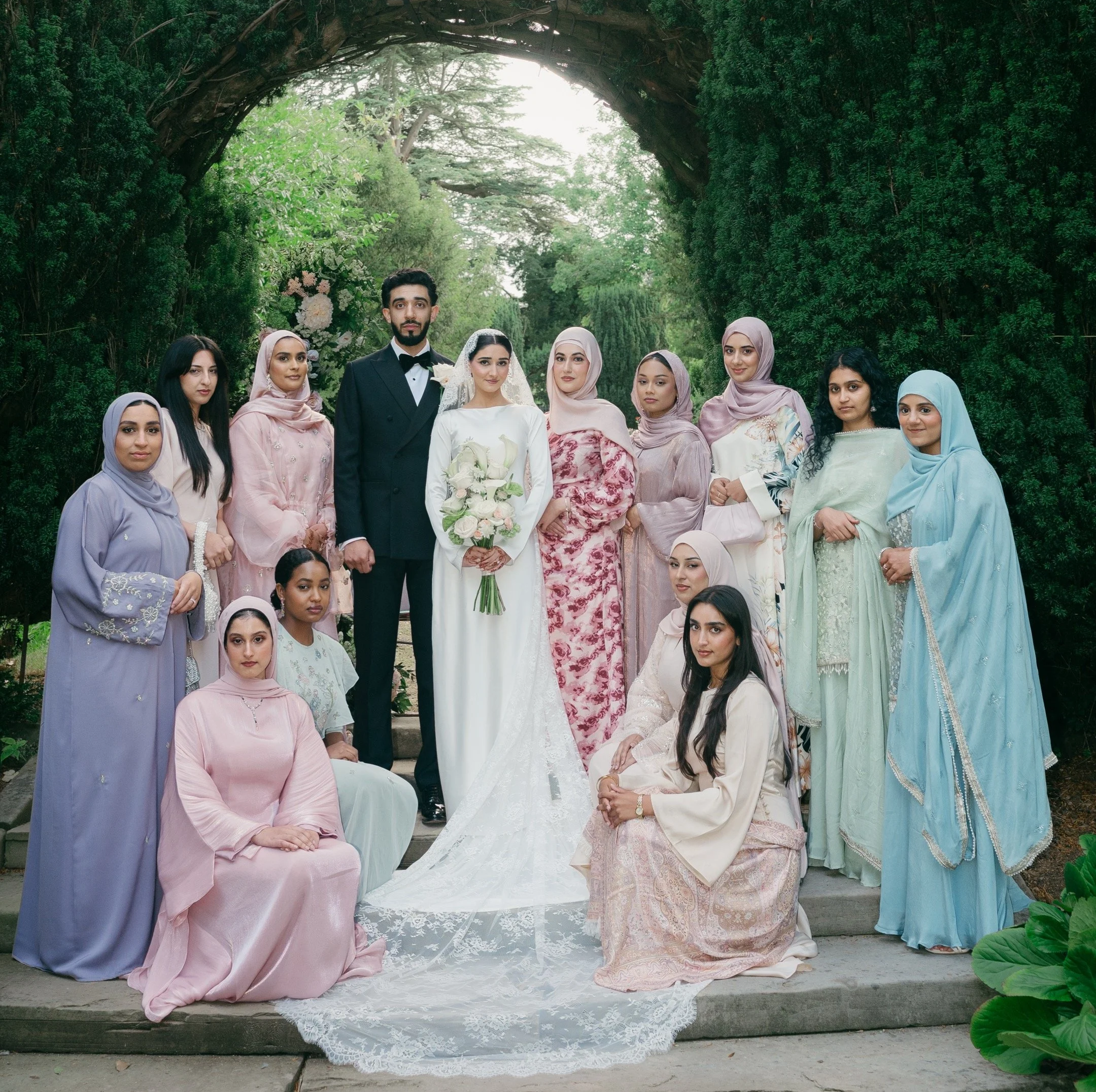 A wedding group photo outdoors featuring a bride in a white gown with a lace veil holding a bouquet of white roses, a groom in a black tuxedo, and fourteen women dressed in pastel and floral traditional attire, including hijabs, standing on stone steps under a large arch of greenery with trees in the background.
