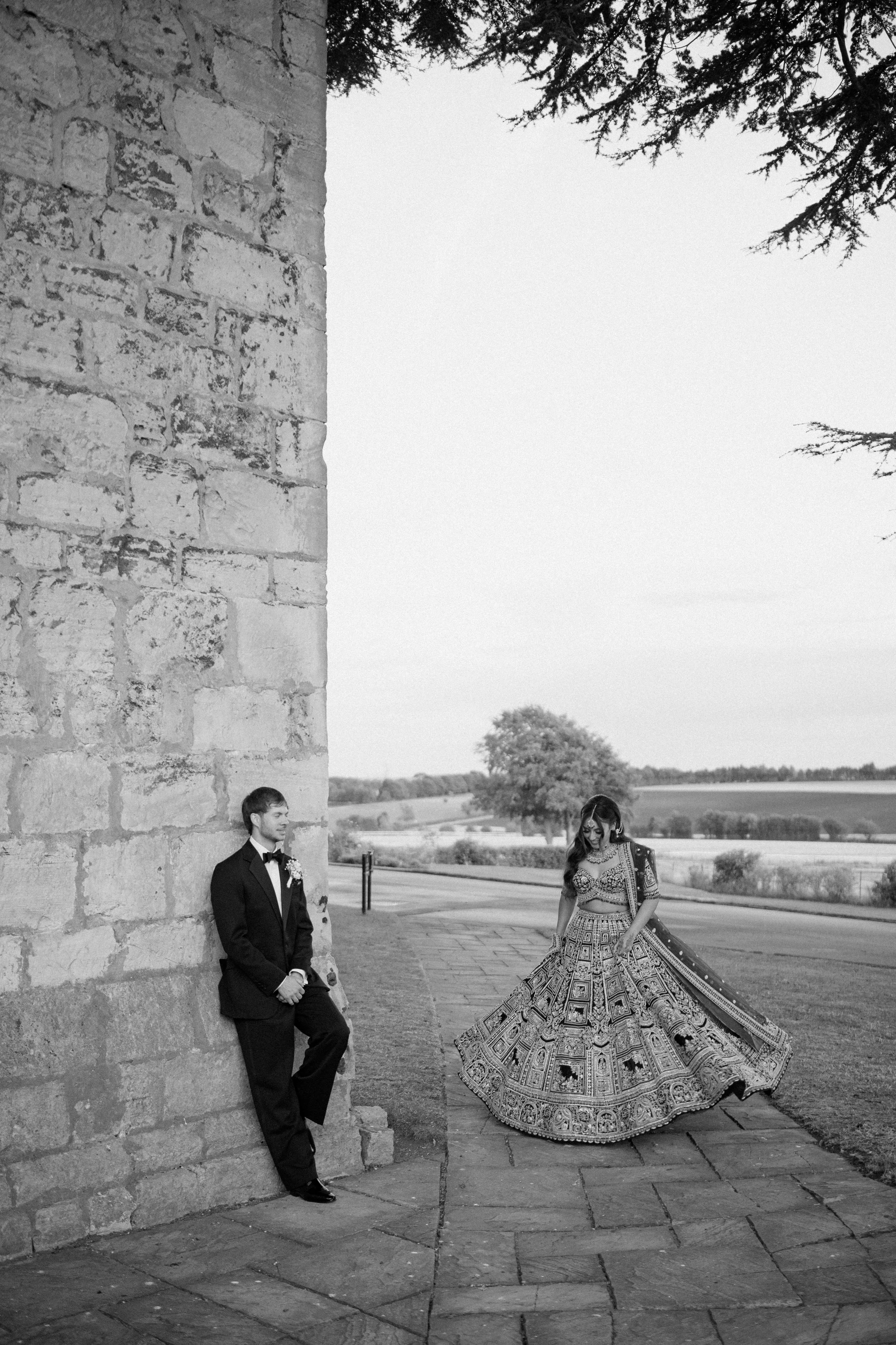 A man in a tuxedo and a woman in traditional Indian attire pose outdoors near a stone wall, with trees and open fields in the background.