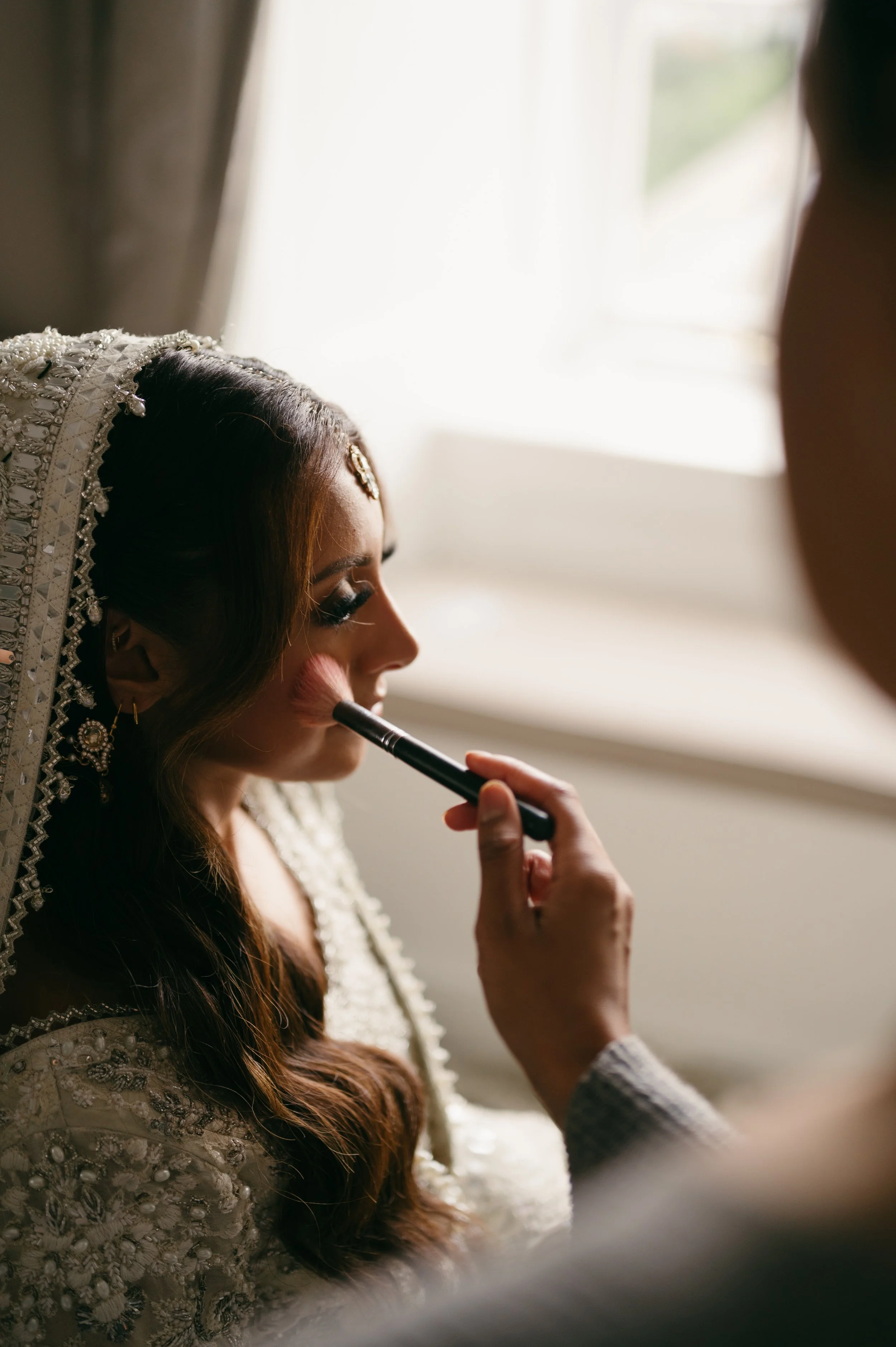 Close-up of a bride with intricate jewelry and a traditional dress, as a makeup artist applies blush to her cheek.