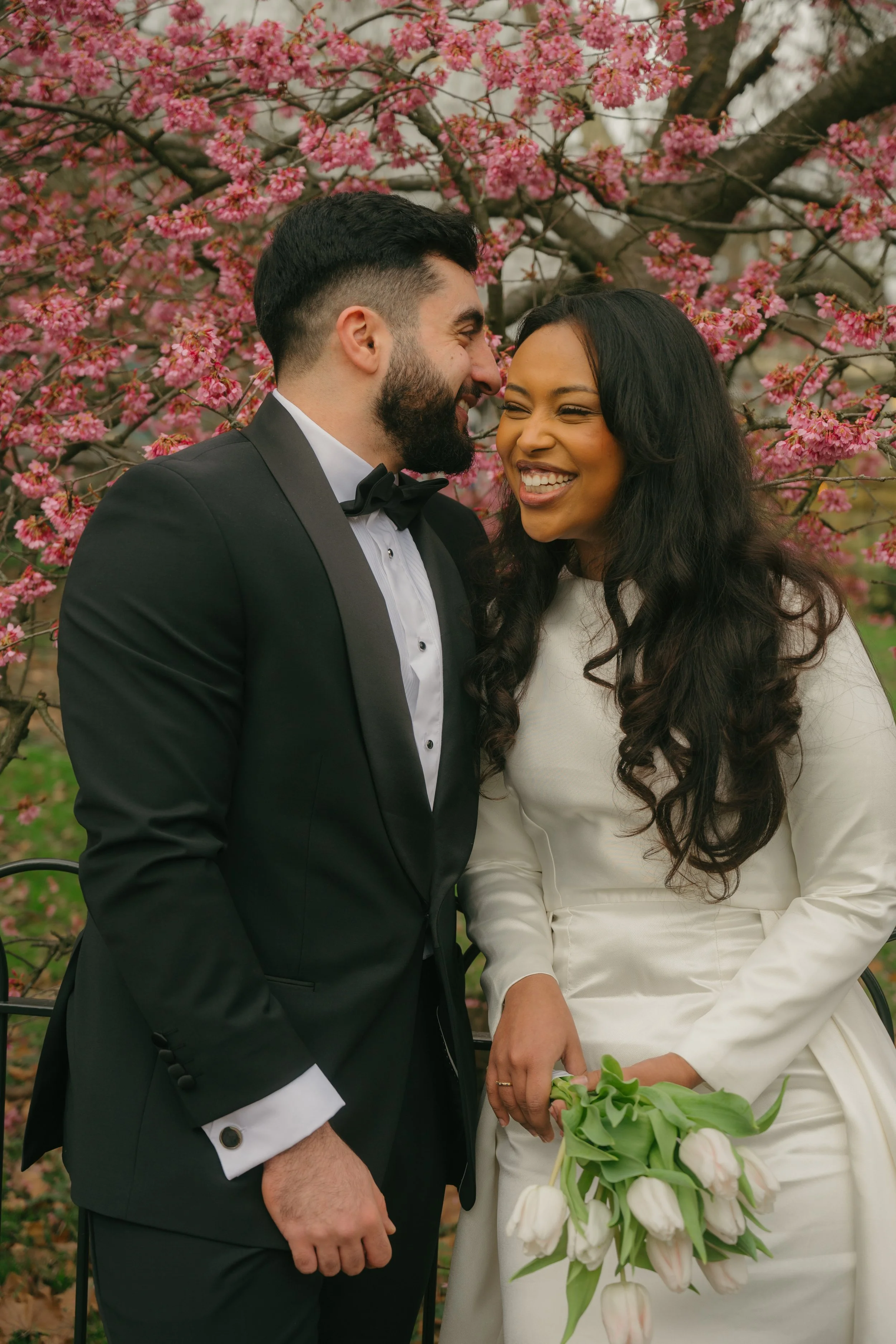 A newlywed couple smiling and leaning close to each other outdoors in front of blooming pink cherry blossom trees. The groom is wearing a black tuxedo with a white shirt and black bow tie. The bride is wearing a white dress, holding a bouquet of whit