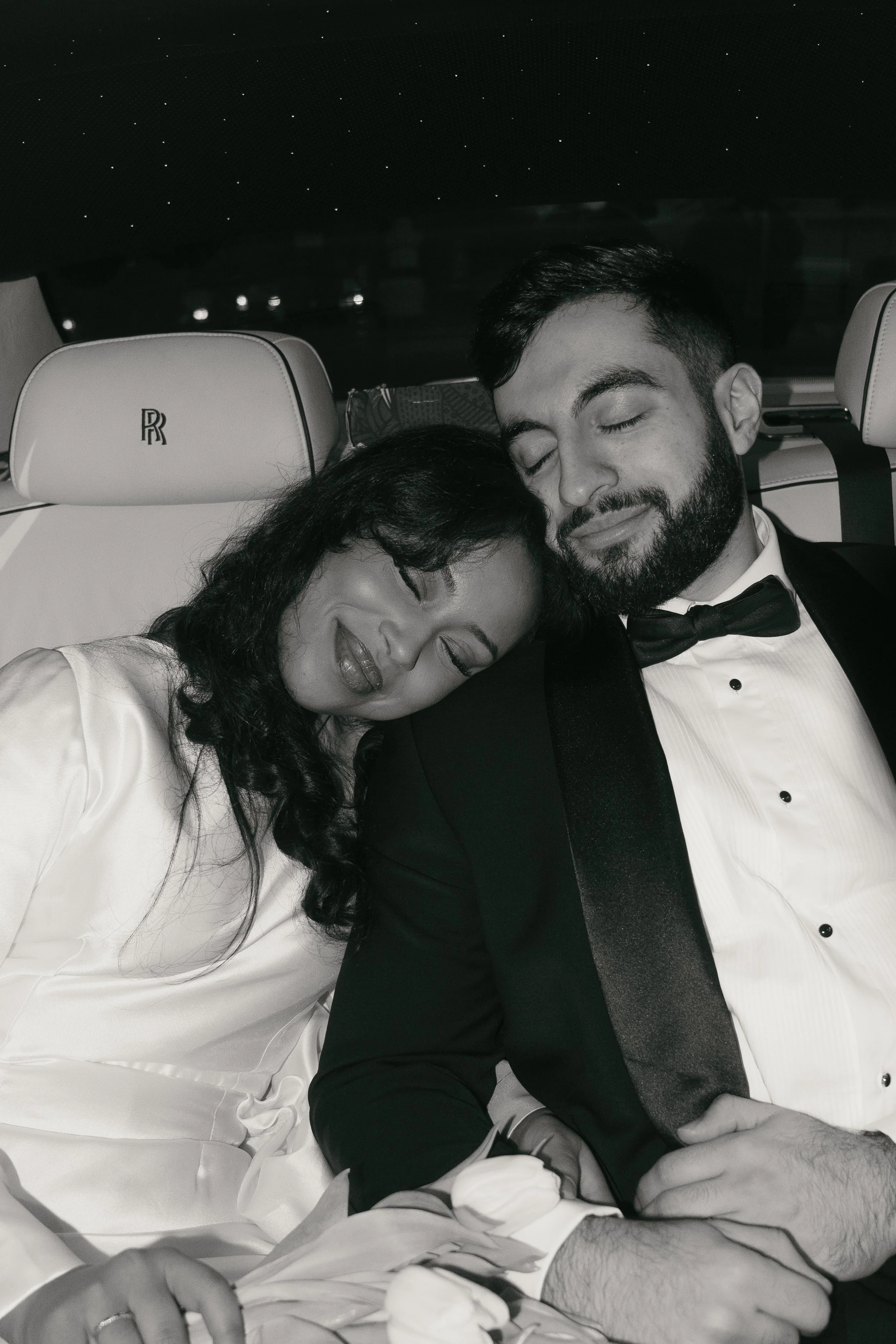 A black and white photo of a couple sleeping in a car, dressed in formal attire. The woman has curly dark hair and is wearing a white dress, holding a bouquet of white flowers. The man has a beard, is wearing a tuxedo with a bow tie, and both have pe