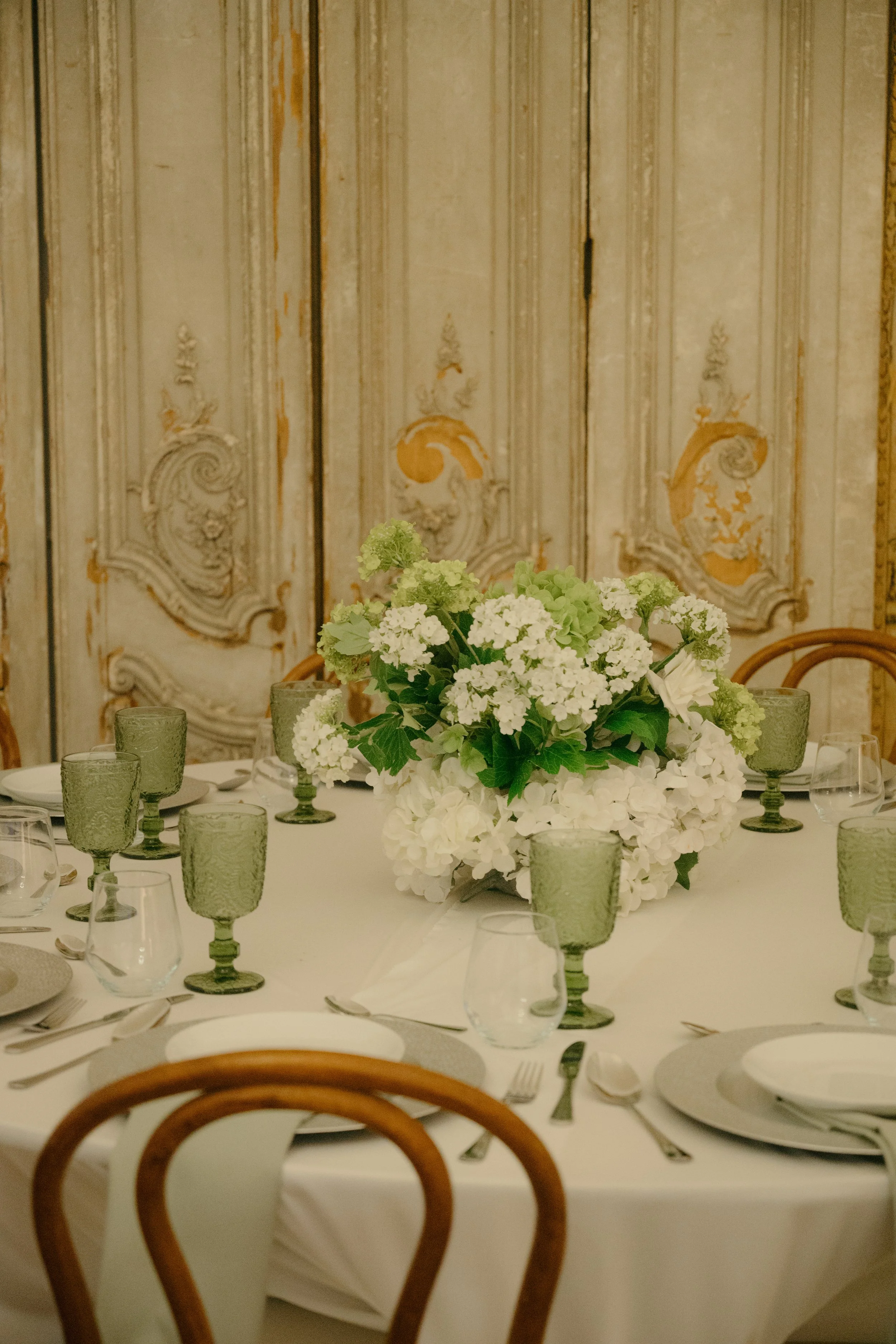 A round dining table decorated with a large white floral centerpiece, surrounded by white plates, silverware, wine glasses, and green goblets, with a carved wooden chair visible in the foreground, and an ornate, vintage wall in the background.