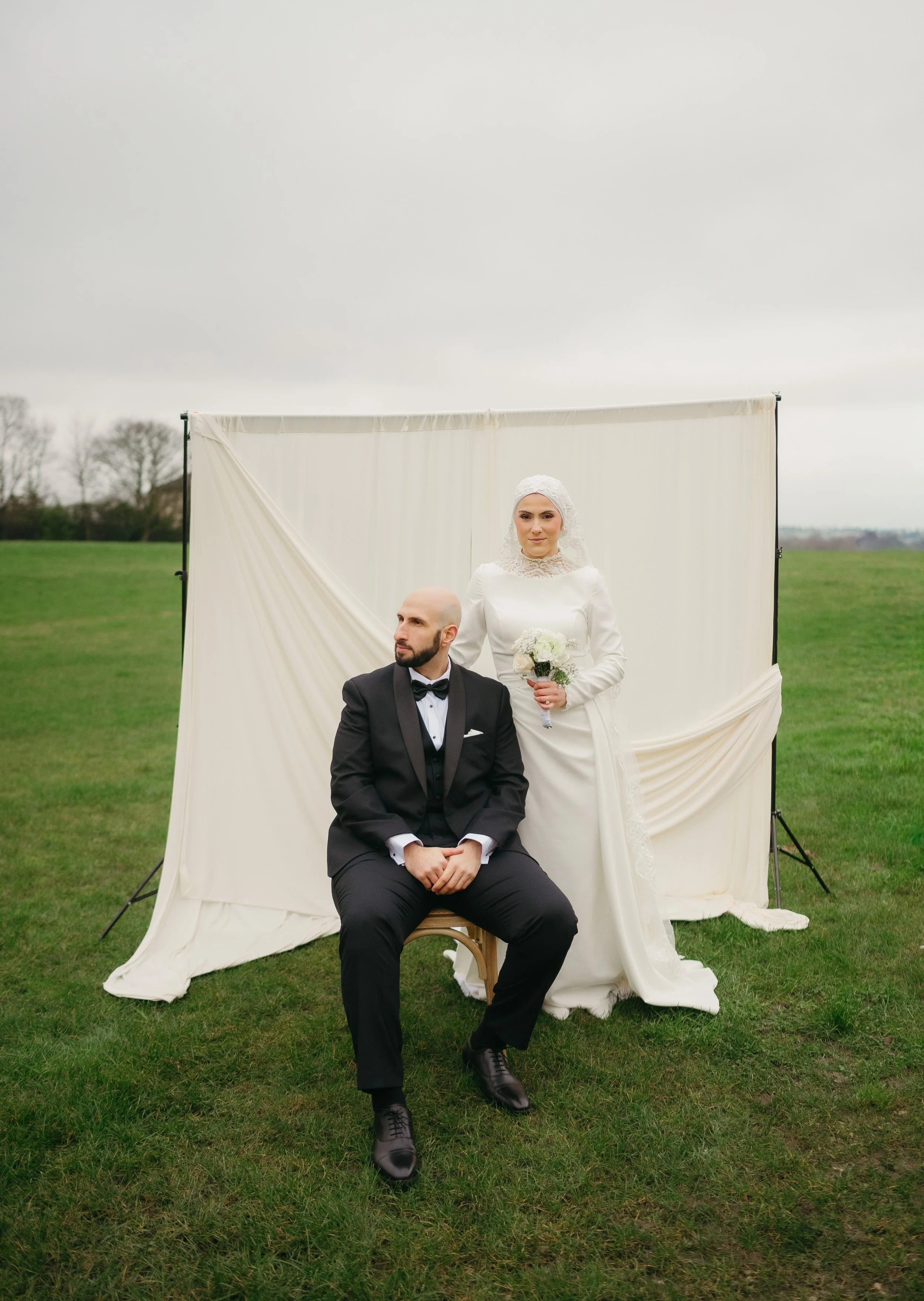 A bride and groom in wedding attire posing outdoors in front of a white fabric backdrop on a grassy field, with a cloudy sky overhead. The groom is seated, wearing a black tuxedo and bow tie, while the bride stands beside him holding a bouquet, dress