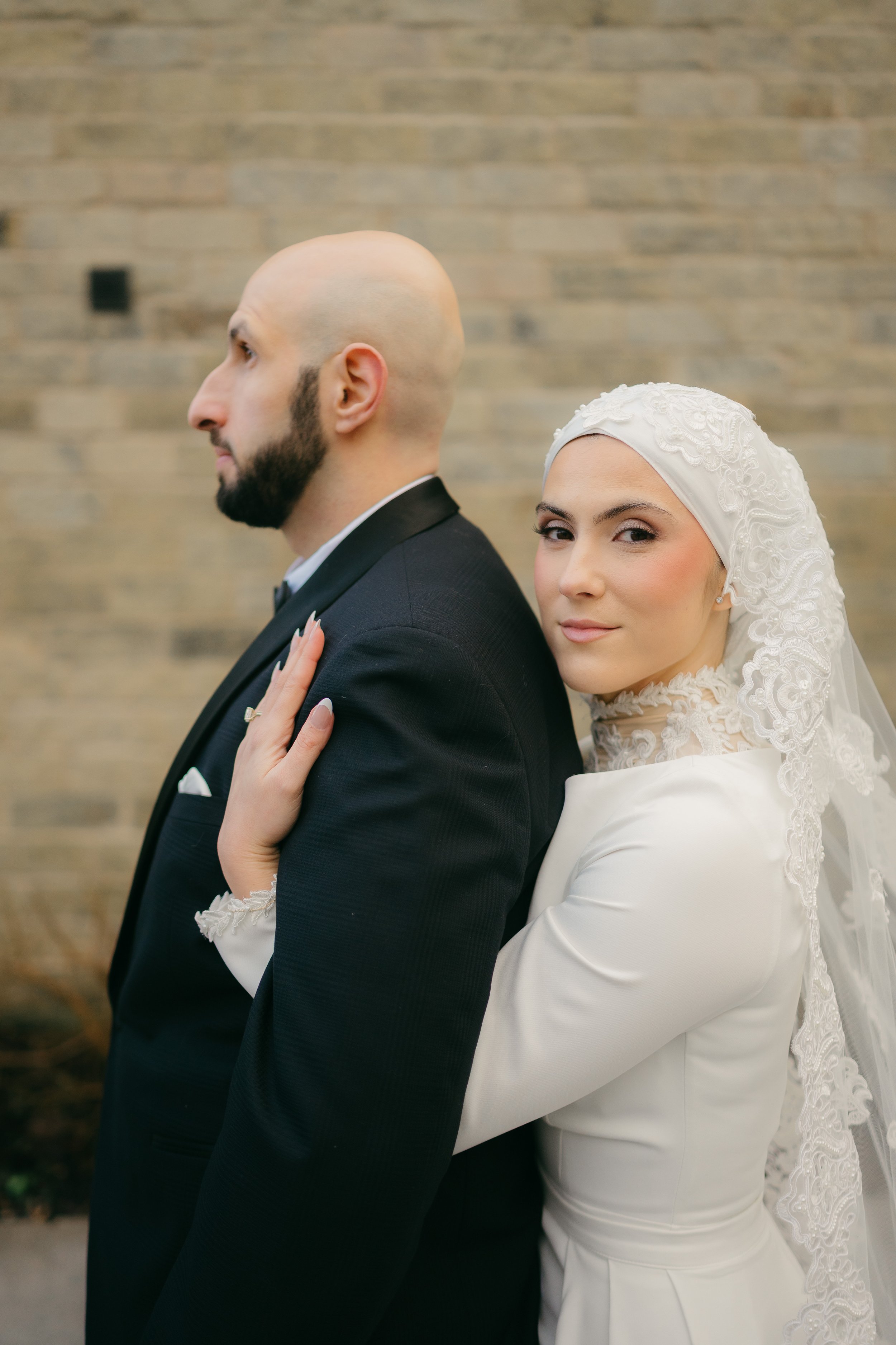 A bride in a white wedding dress and lace hijab hugging a groom in a black tuxedo with a bald head and beard, standing in front of a brick wall.