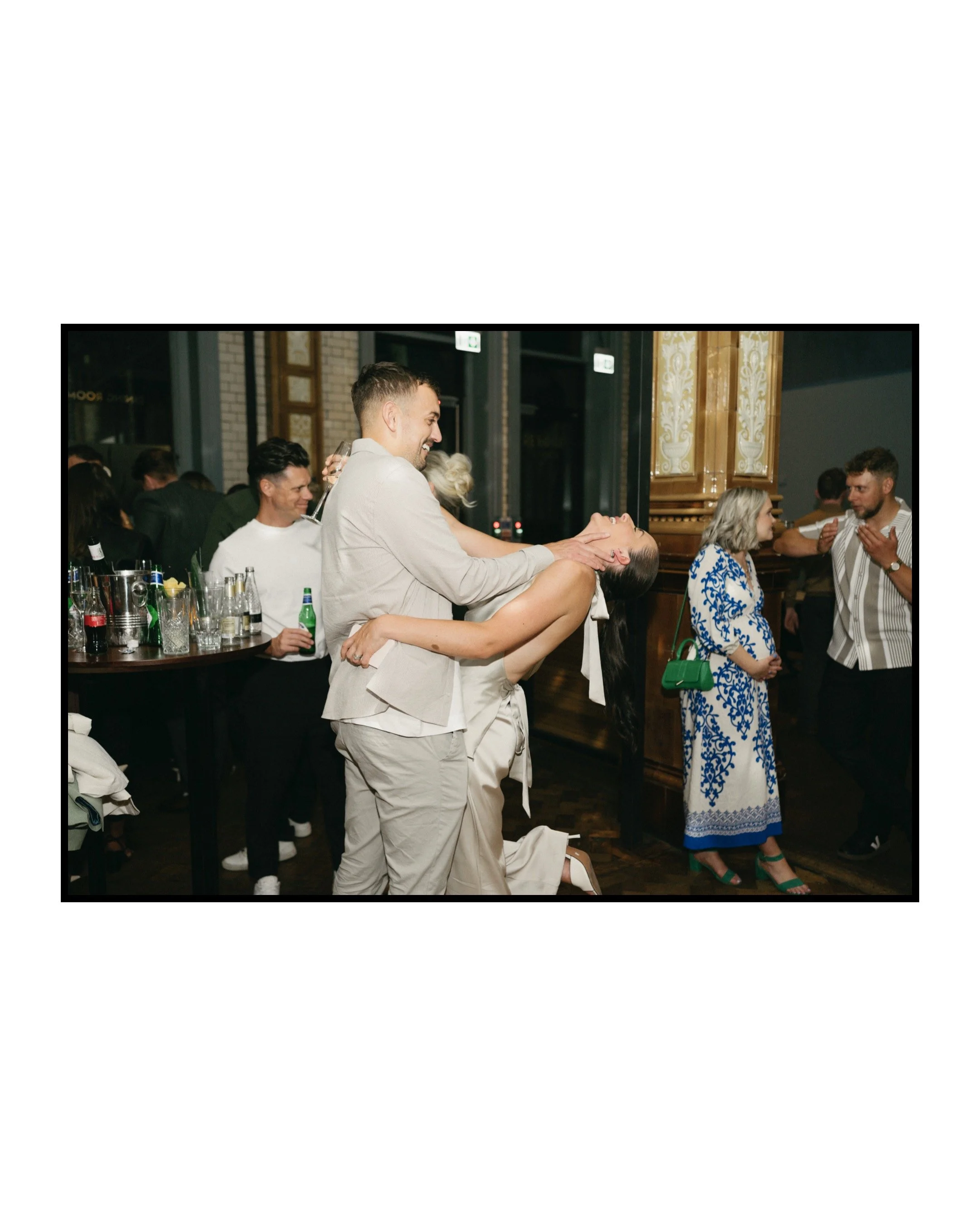 A group of people celebrating at a party, with a man dipping a woman and both smiling, while other guests converse and enjoy drinks in the background.