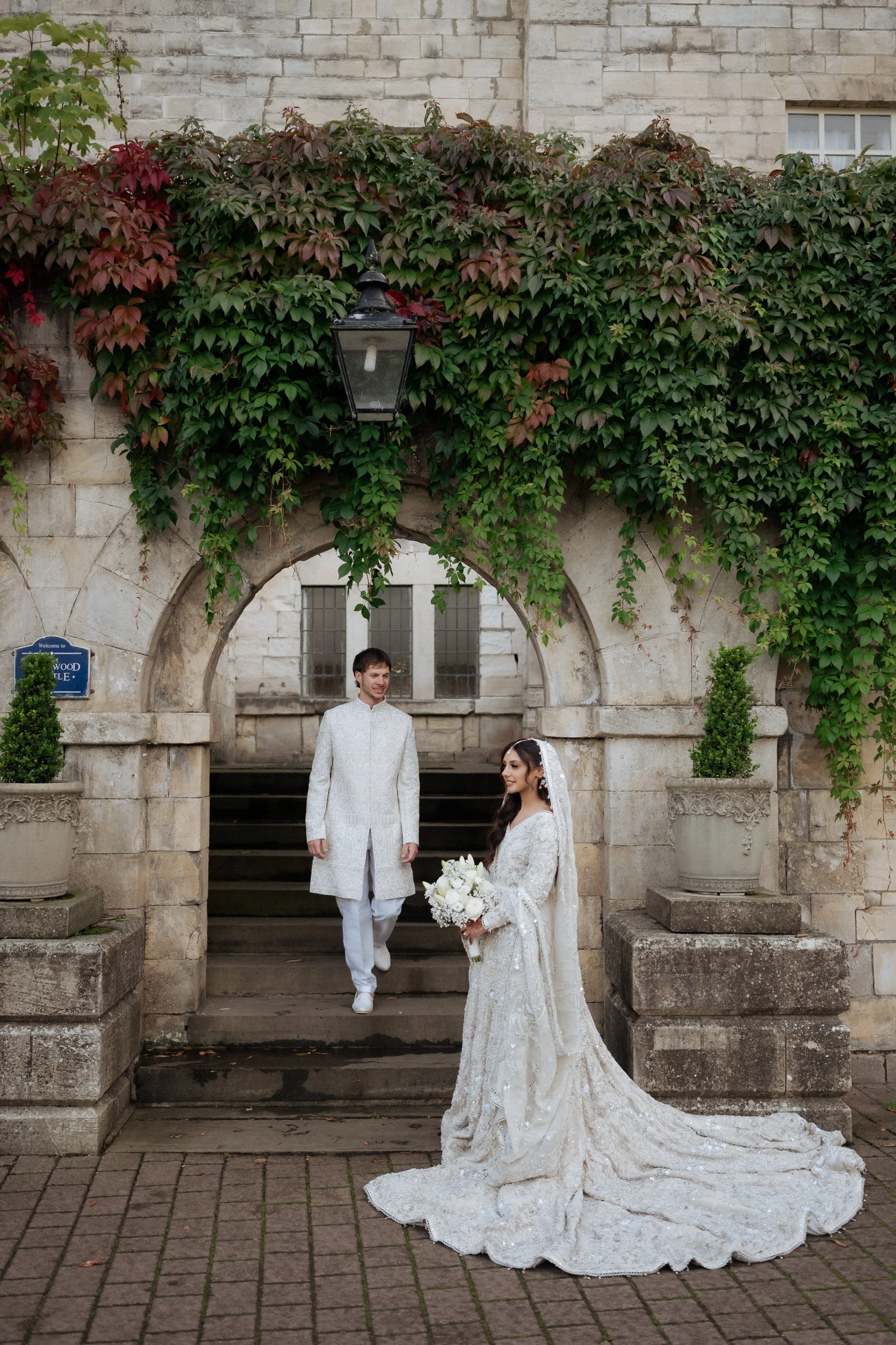 A bride and groom on their wedding day, standing outside under an arch covered in green and red ivy, with brick and stone architecture in the background.