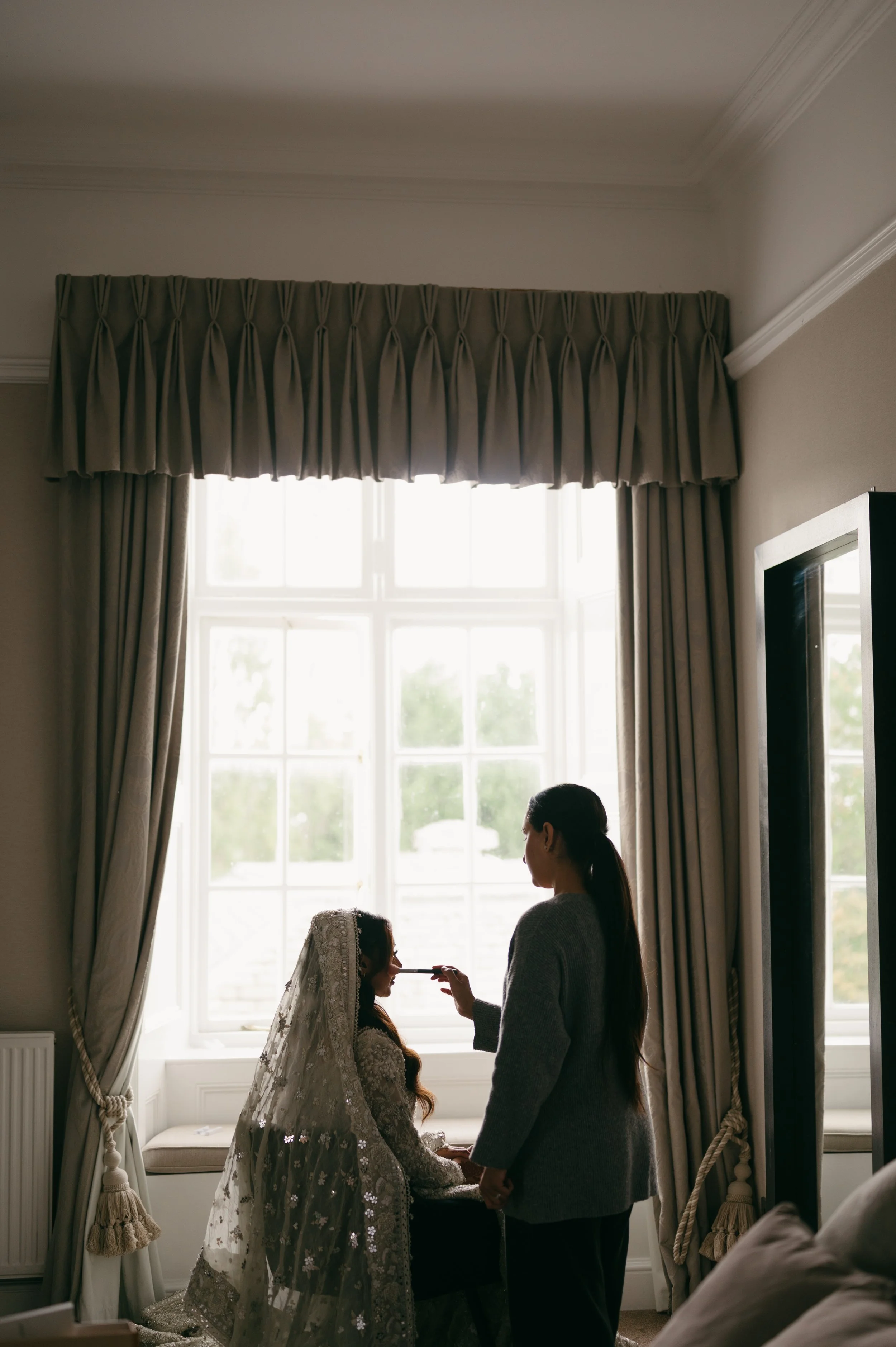 A woman puts makeup on another woman dressed in a wedding gown, sitting by a window with curtains.