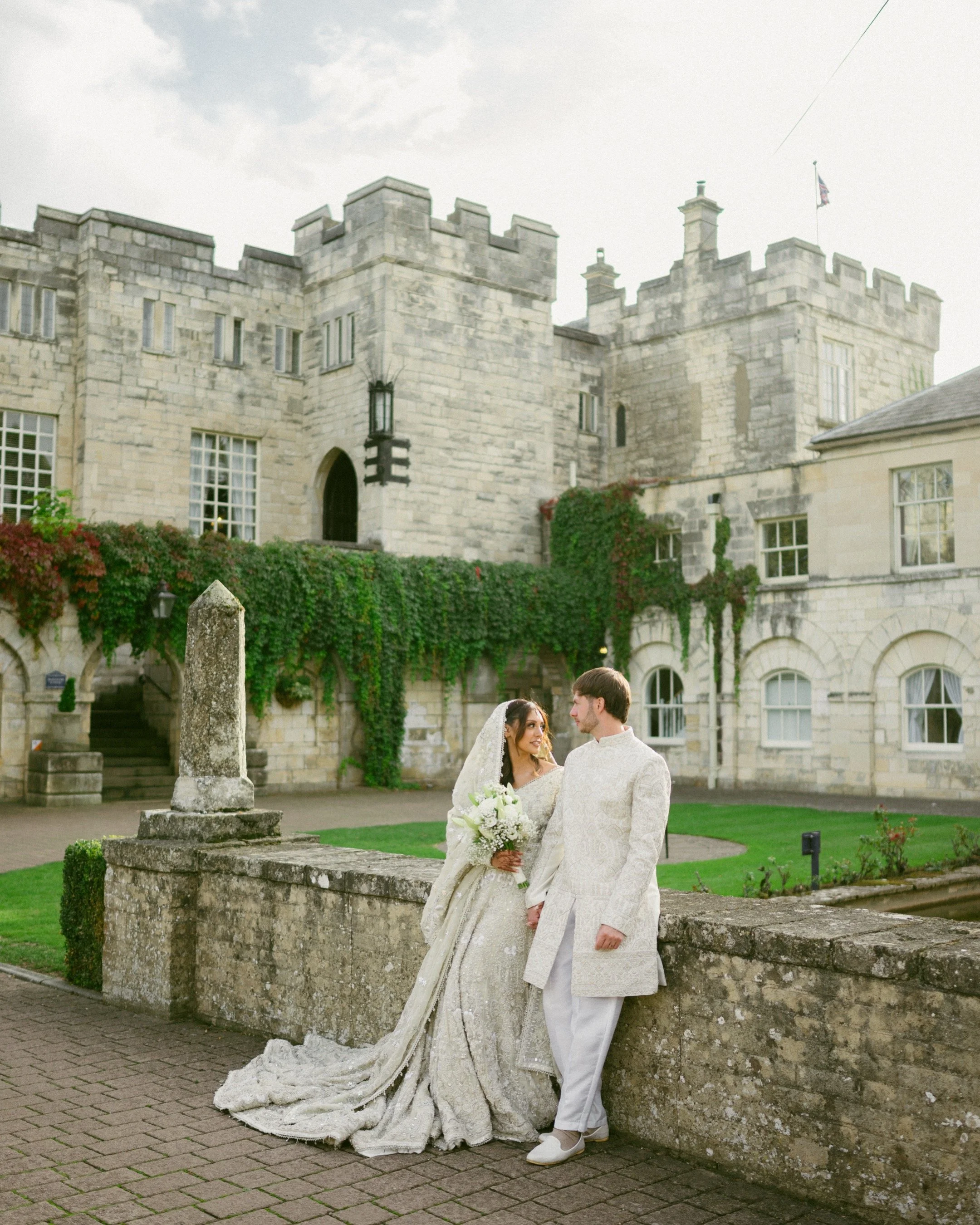 A bride and groom in wedding attire standing outside a historic stone castle, holding hands, with the bride holding a bouquet of flowers and looking at the groom.