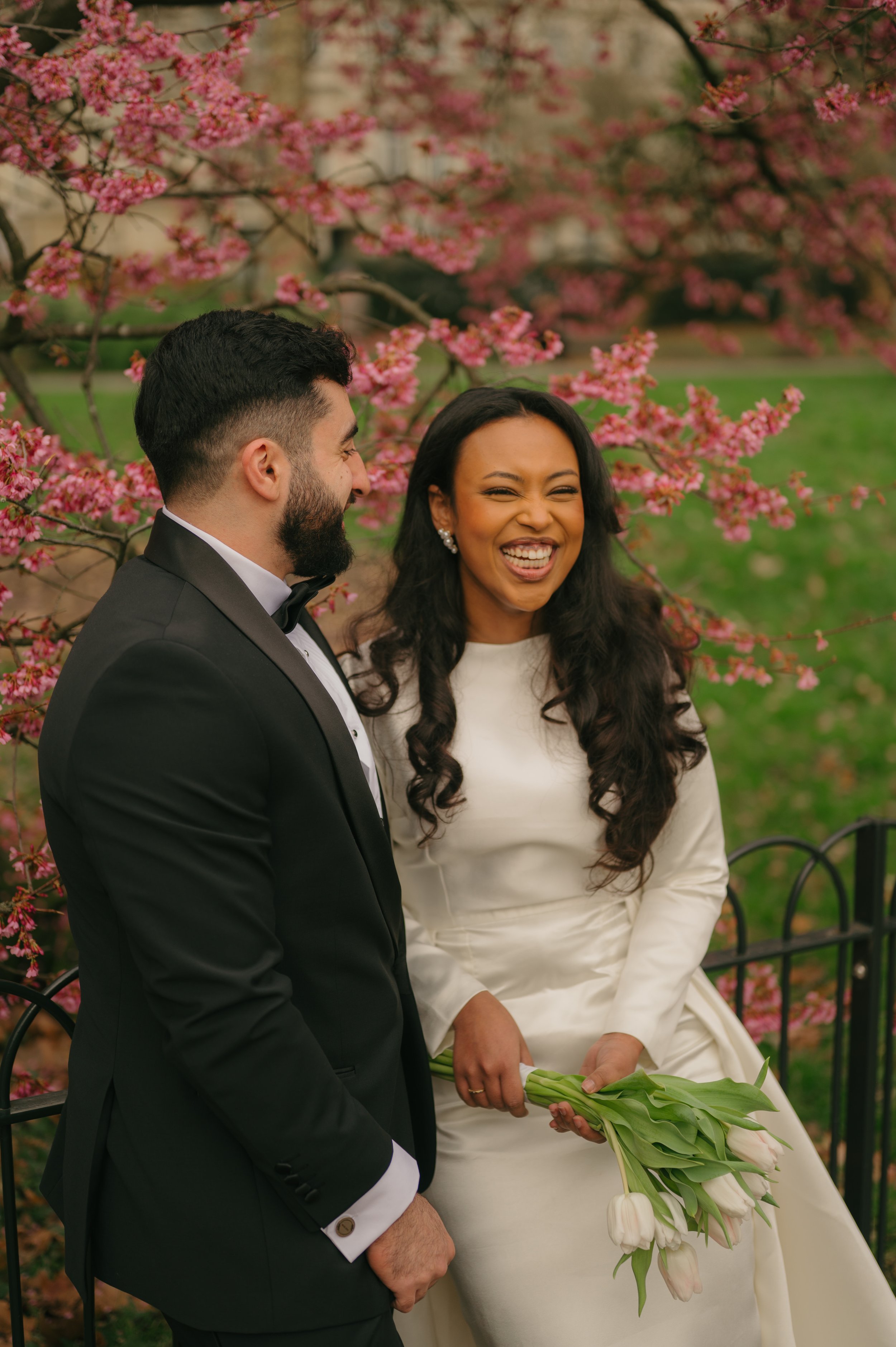 A bride and groom smiling and laughing in a garden with pink blossoms, the bride holding a bouquet of white tulips.