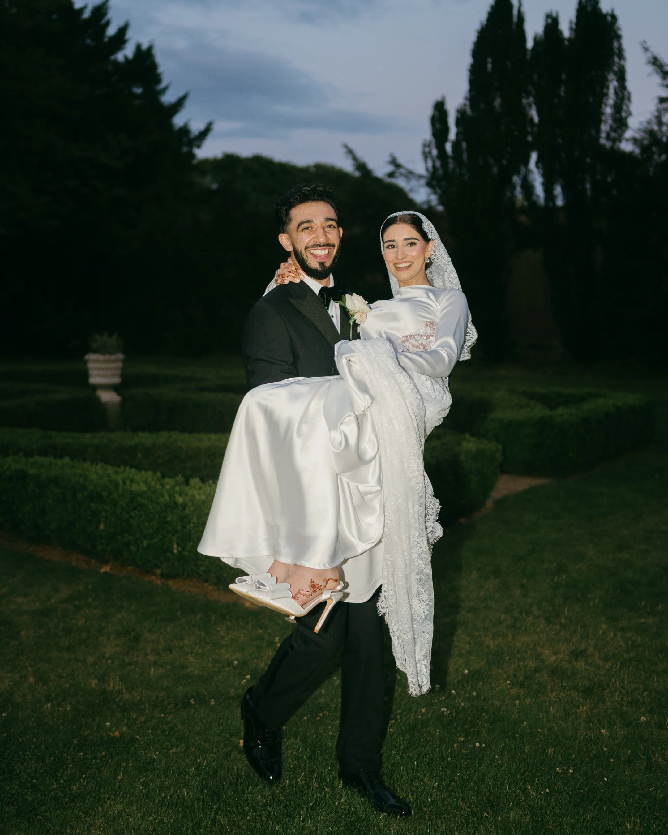 A groom in a black tuxedo carrying a bride in a white wedding dress and veil, outdoors on a lawn during evening.