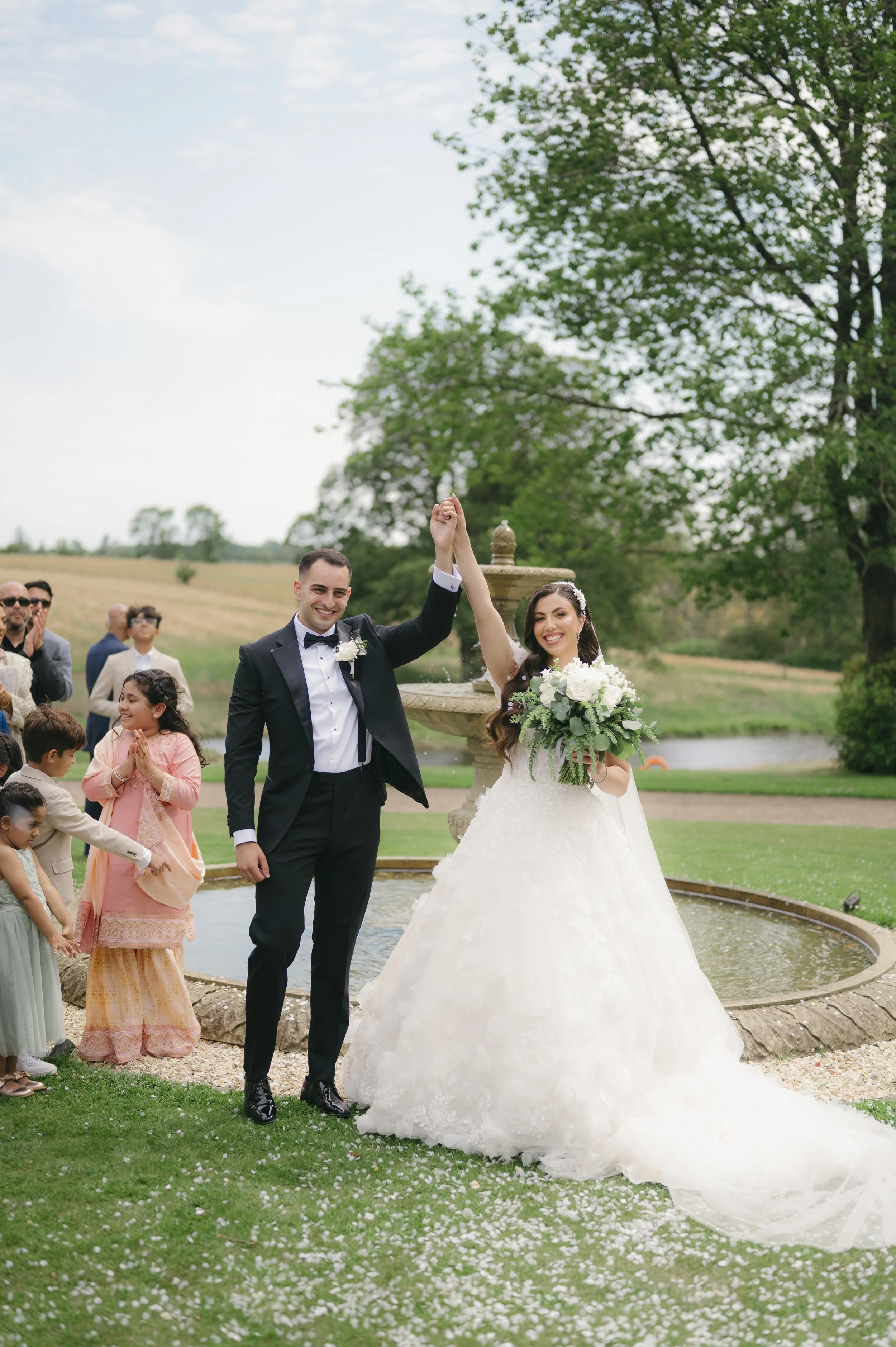 A newlywed couple celebrates outside by a water fountain, with the bride holding a bouquet, as guests rejoice in the background.