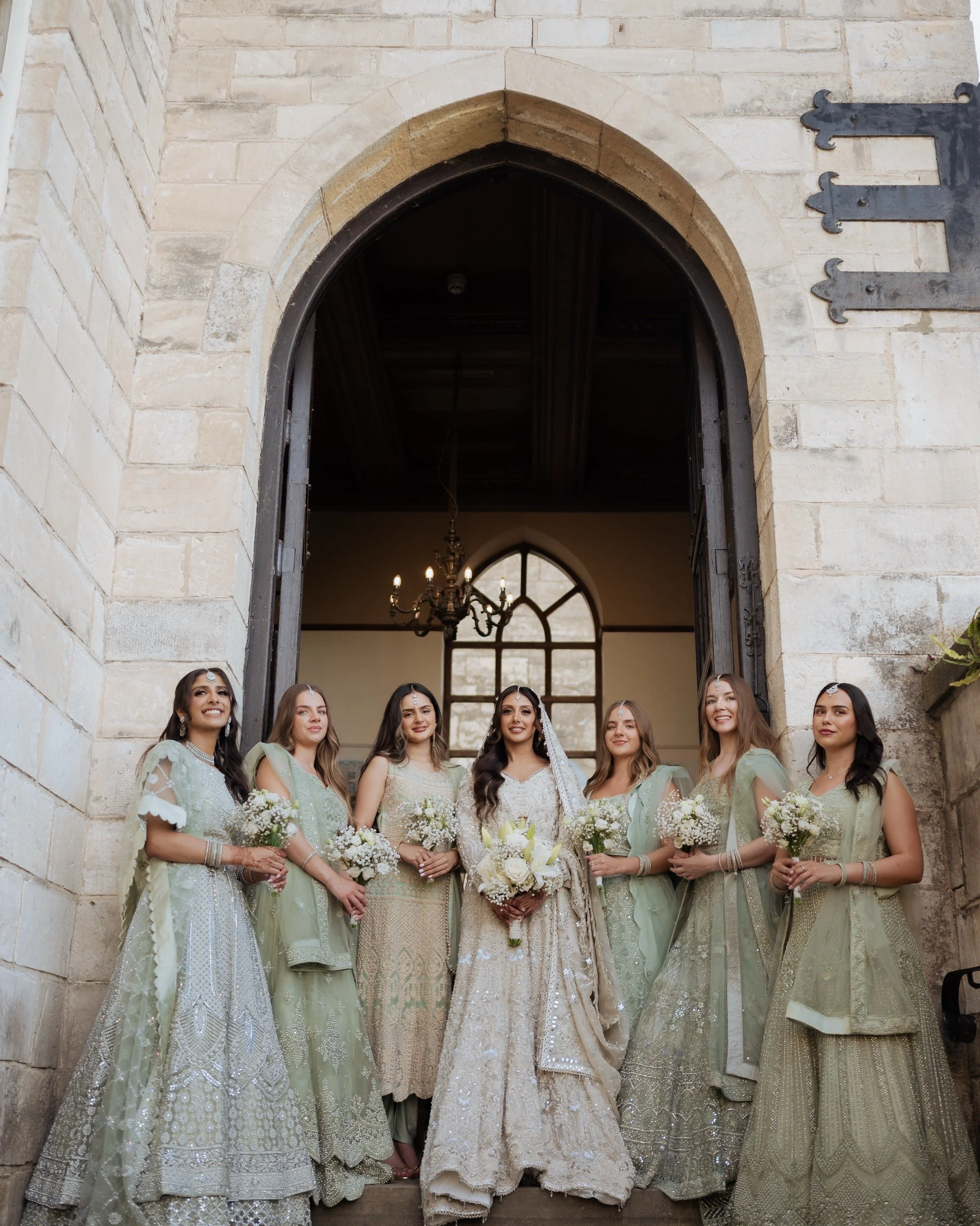 Bride in a white wedding dress and six bridesmaids in matching pastel green and gold outfits standing in a church doorway, holding bouquets for a wedding photoshoot.