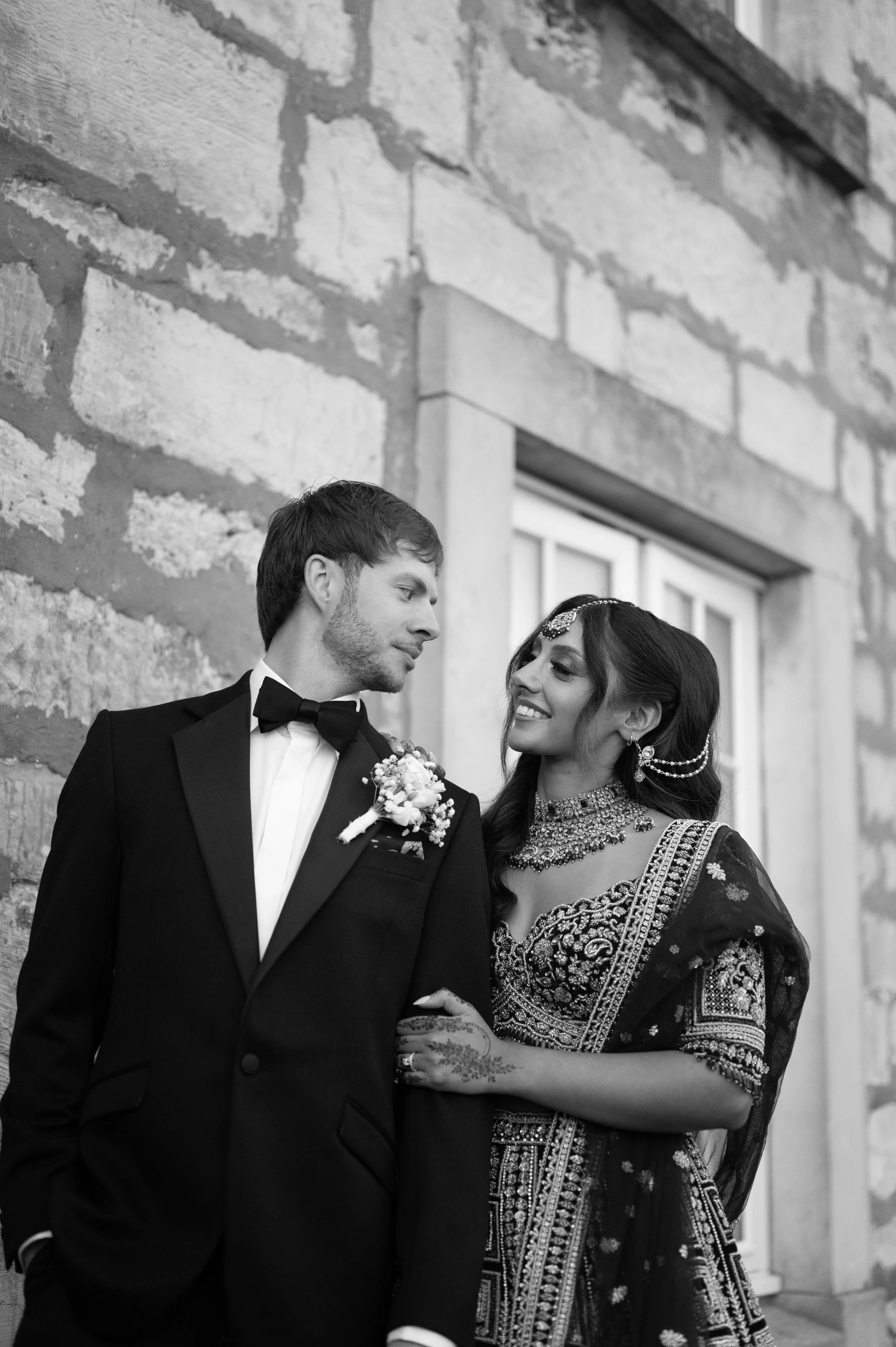 A black and white photo of a couple outdoors against a brick wall. The man is wearing a tuxedo with a bow tie and boutonnière, looking at the woman. The woman is dressed in traditional South Asian attire with jewelry, smiling, and looking at the man,