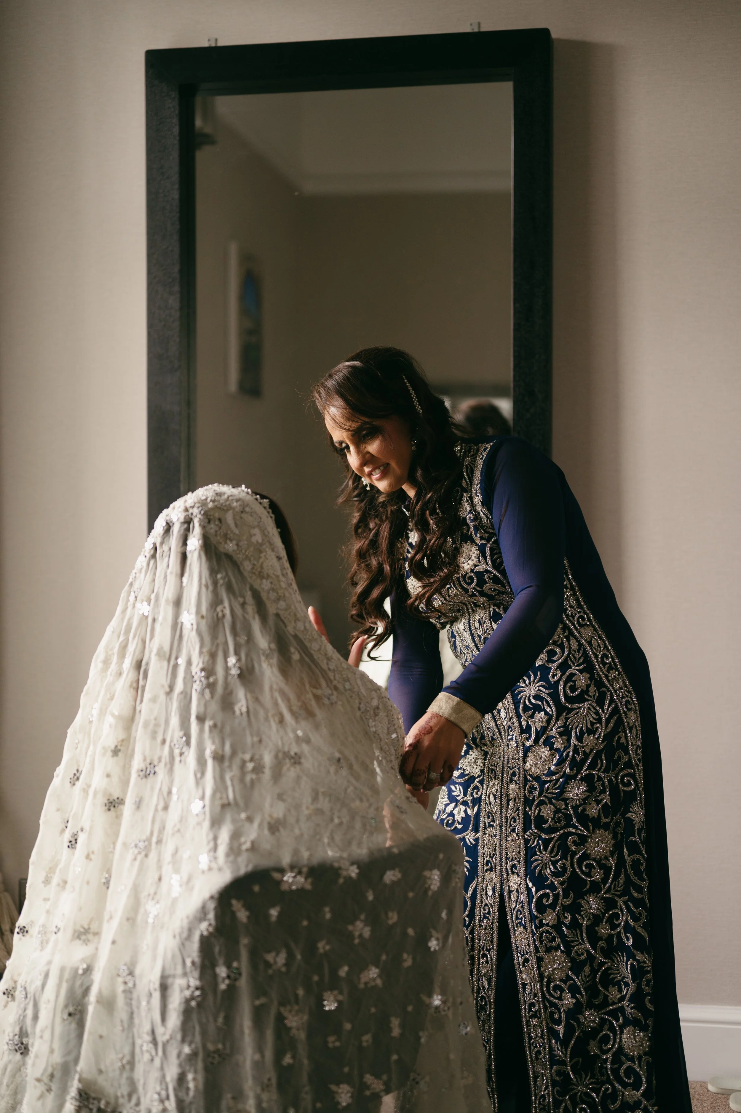 A woman in a blue embroidered dress helping a bride adjust her wedding dress in front of a mirror.
