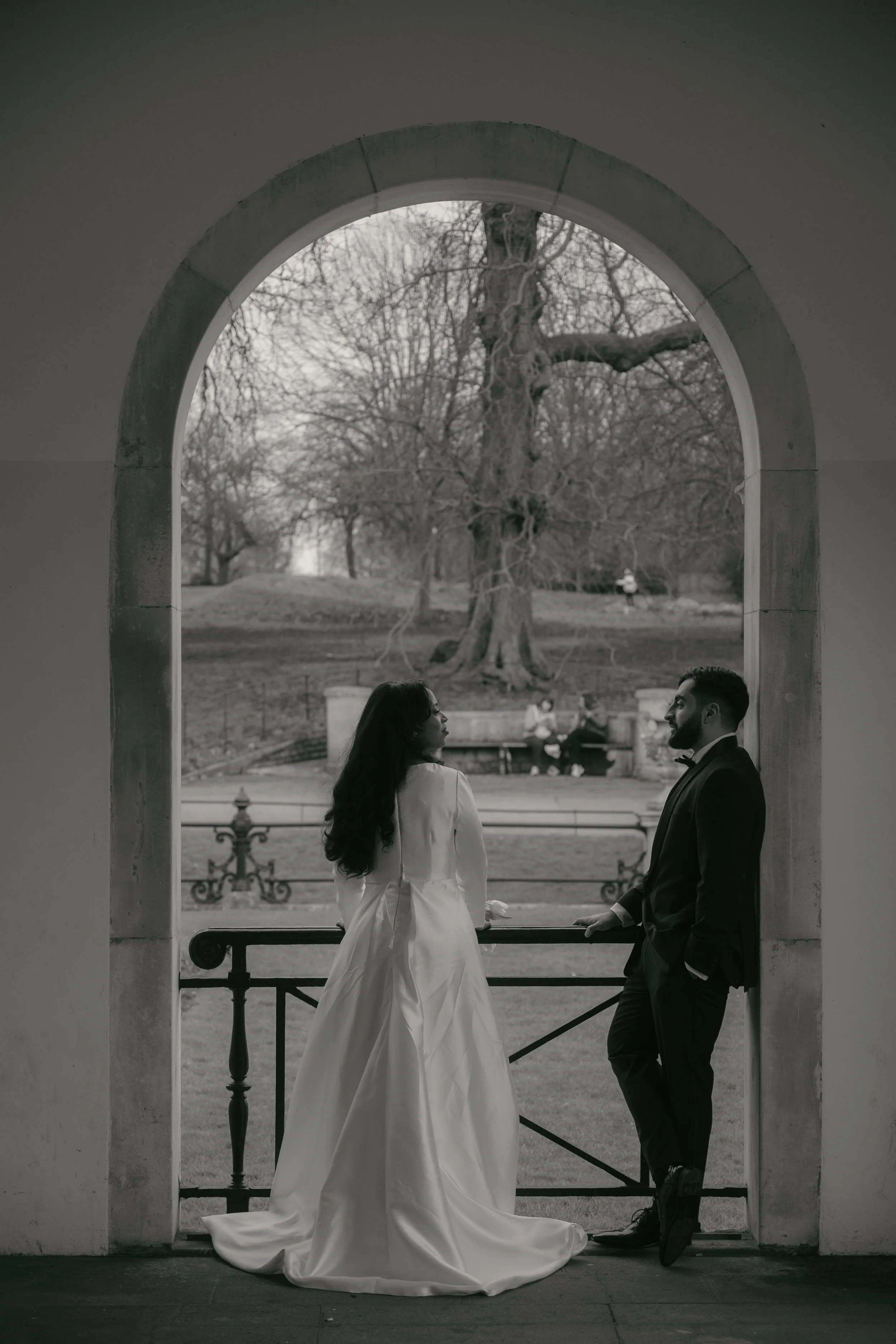 A black and white photograph of a bride and groom standing under an archway in a park. The bride is wearing a long wedding gown, and the groom is in a tuxedo. They are facing each other, with the groom leaning against the archway and the bride lookin