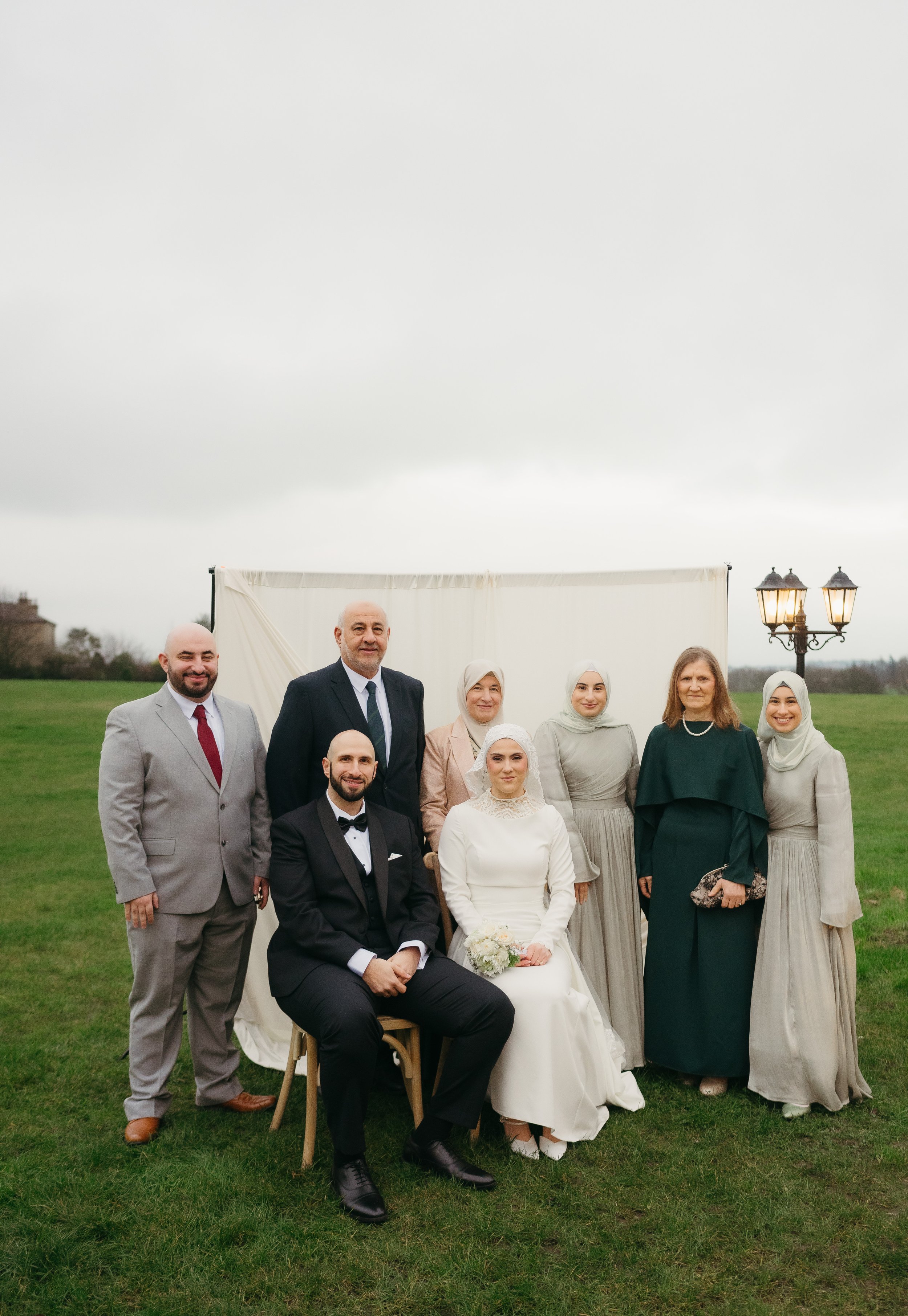 Group of people at an outdoor wedding ceremony, including a bride in a white dress holding a bouquet, a groom in a tuxedo, and six women and one man dressed in formal attire, standing on a grassy field near a white backdrop and vintage street lamp.