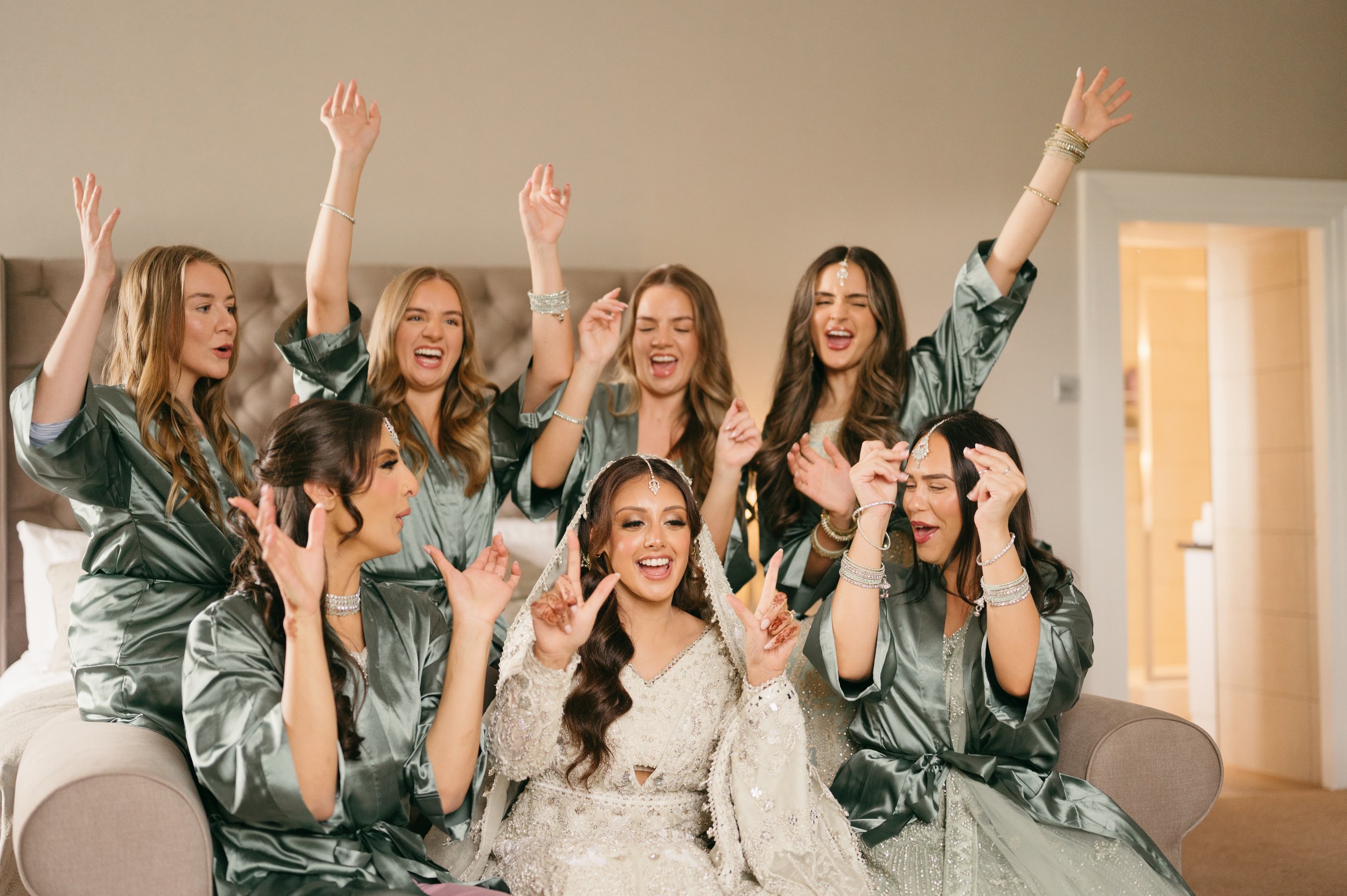 Bridal party celebrating and dancing in a room, with bridesmaids in matching green robes and the bride in white, all smiling and making joyful gestures.